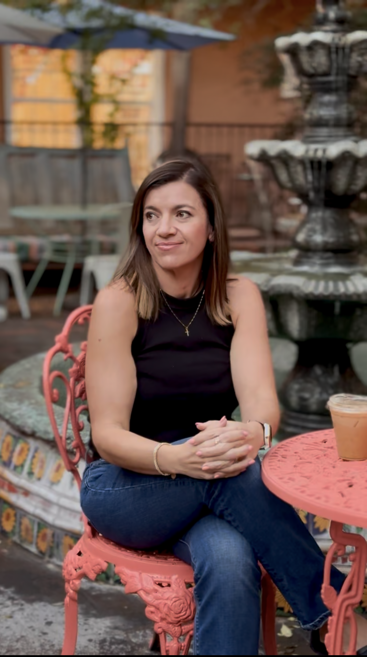 A woman sitting at a pink decorative outdoor table with a carved floral pattern, in a garden or backyard setting with a multi-tiered fountain, a metal chair, and a fence in the background, during late afternoon or early evening.