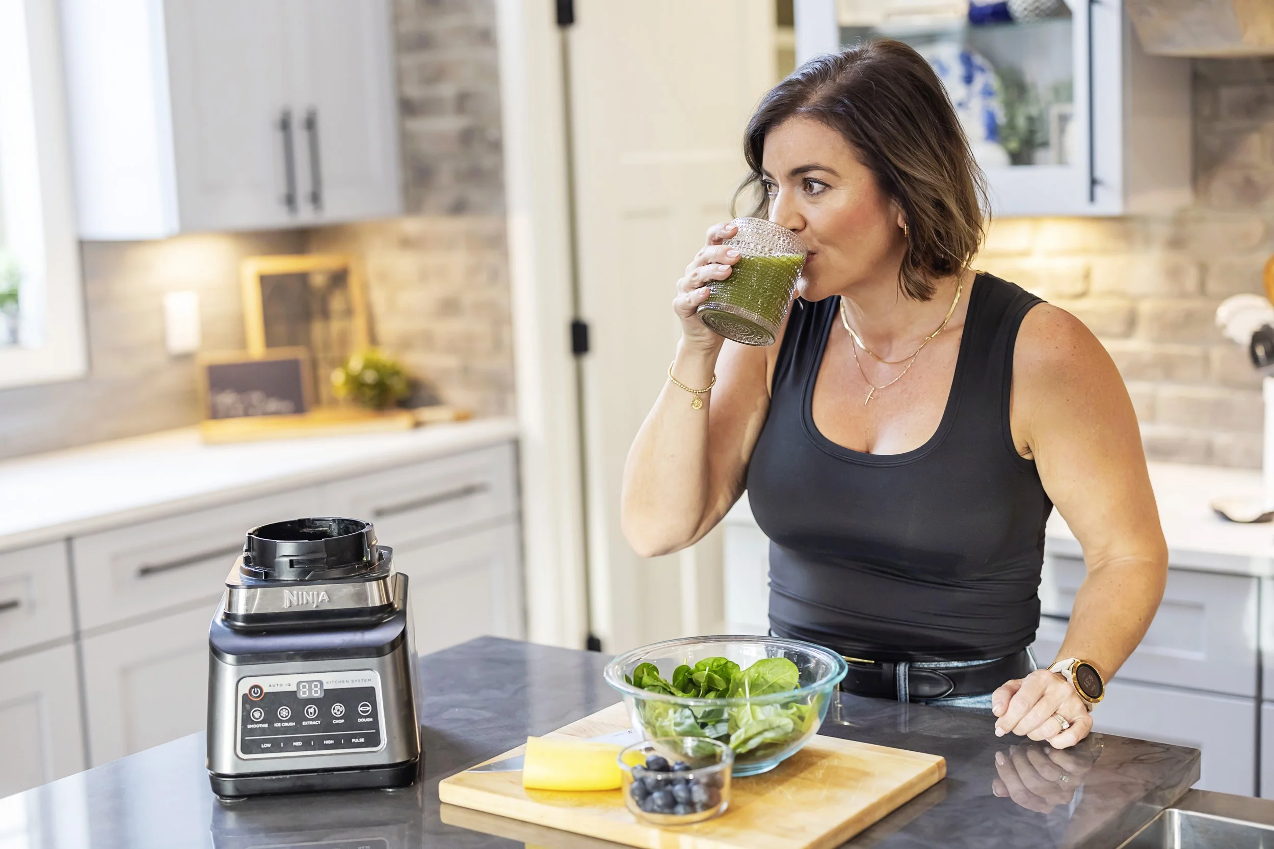 Woman drinking green smoothie in a kitchen with a blender, salad bowl, and blueberries on the counter.