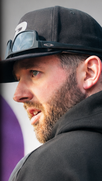 Side profile of a man wearing a black baseball cap, sunglasses on the cap, and a dark hoodie.