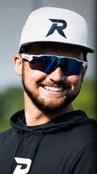 Young man smiling outdoors wearing a white baseball cap with a black logo and reflective sunglasses, dressed in a black hoodie.