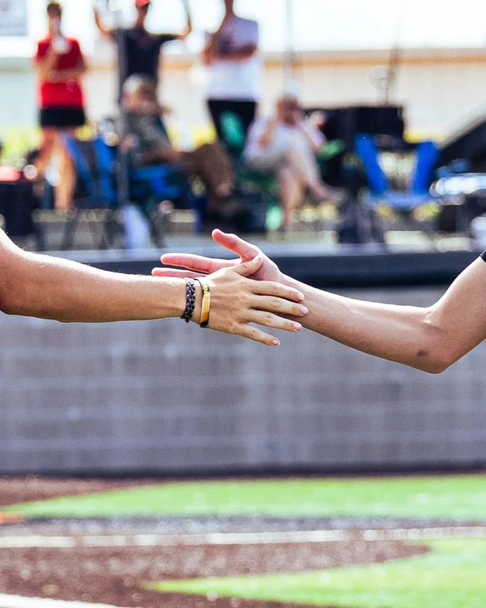 Two people reaching out to shake hands at a baseball or softball field with blurred spectators in the background.