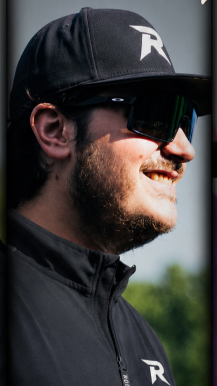 Close-up of a man wearing a black cap with a white logo, reflective sports sunglasses, and a black zip-up jacket with a logo on the chest, smiling outdoors.