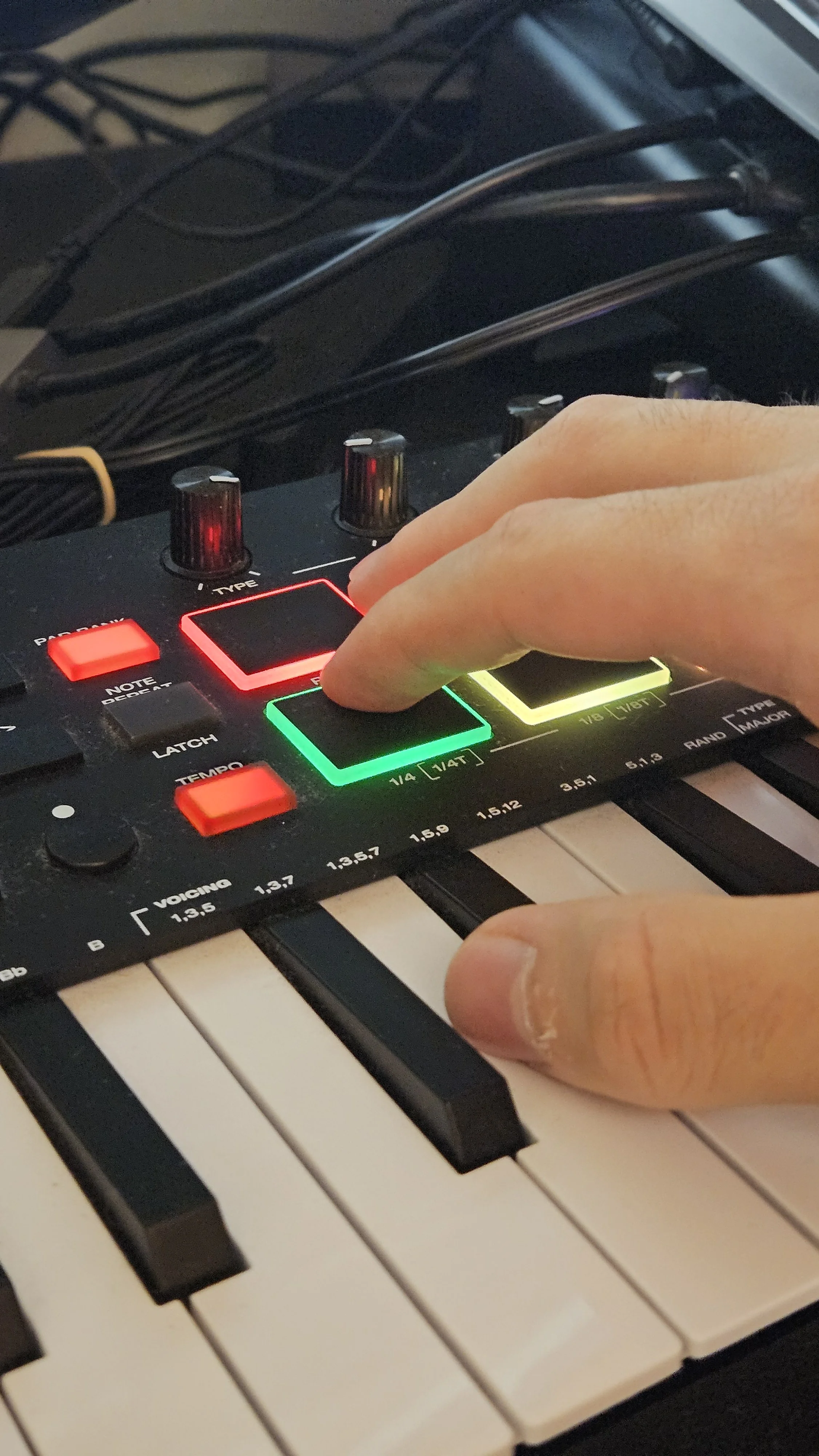 Close-up of a person's hand adjusting controls on a music synthesizer or keyboard, with multiple cables visible in the background.