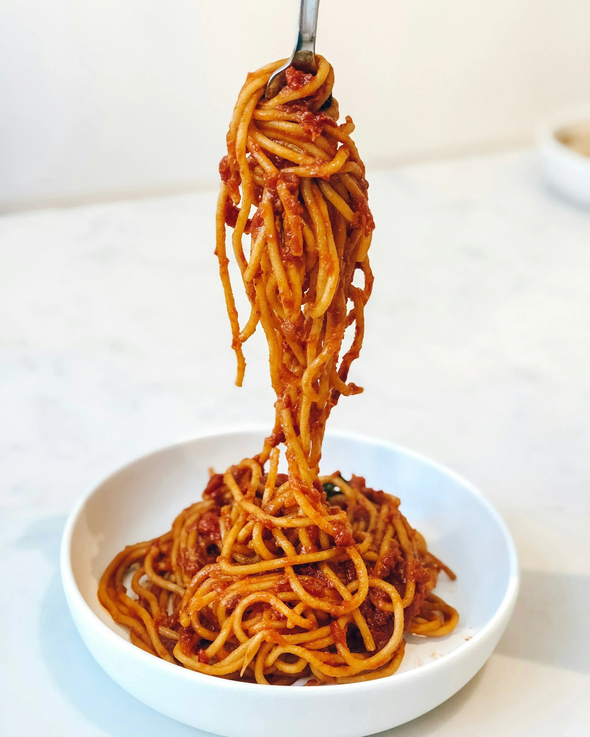 Close-up of a spoon lifting a serving of spaghetti with tomato sauce from a white bowl.