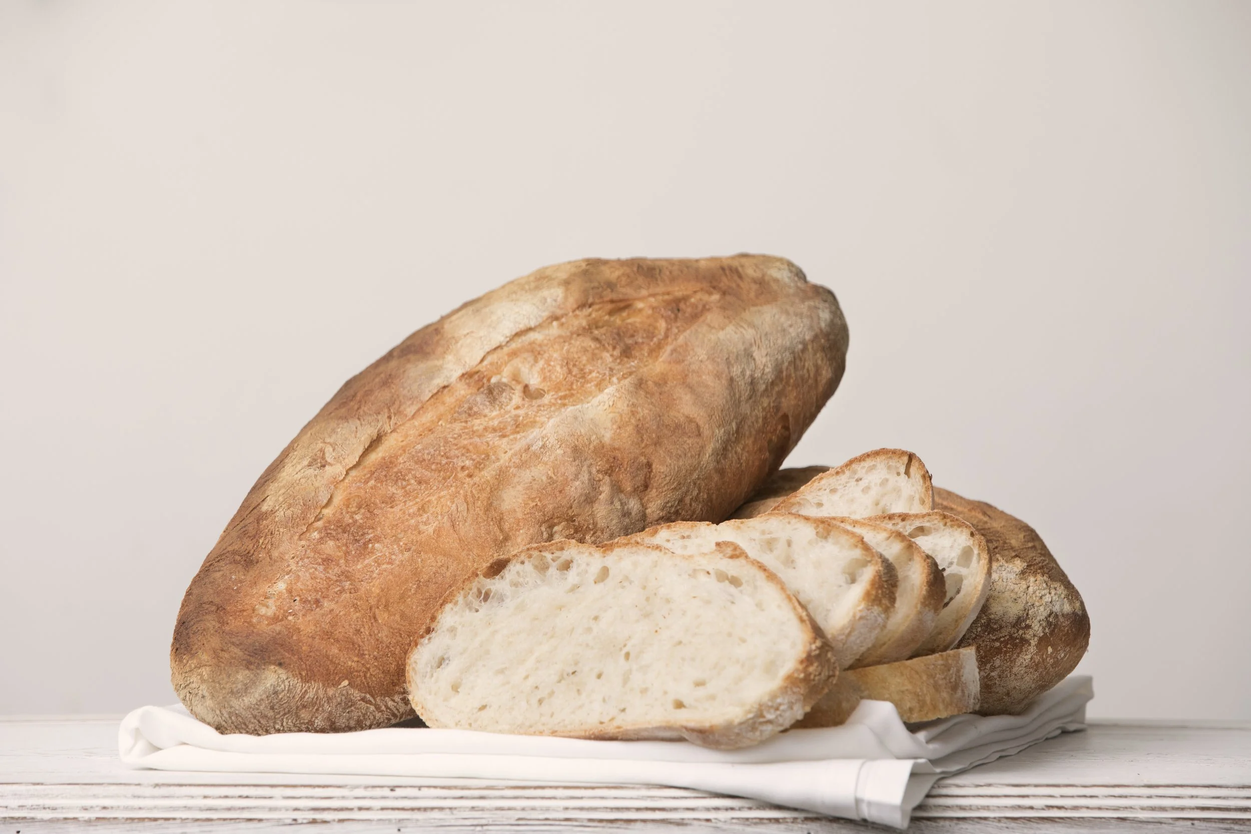Sliced and unsliced loaf of rustic bread on a white plate with a white napkin, on a white wooden surface.
