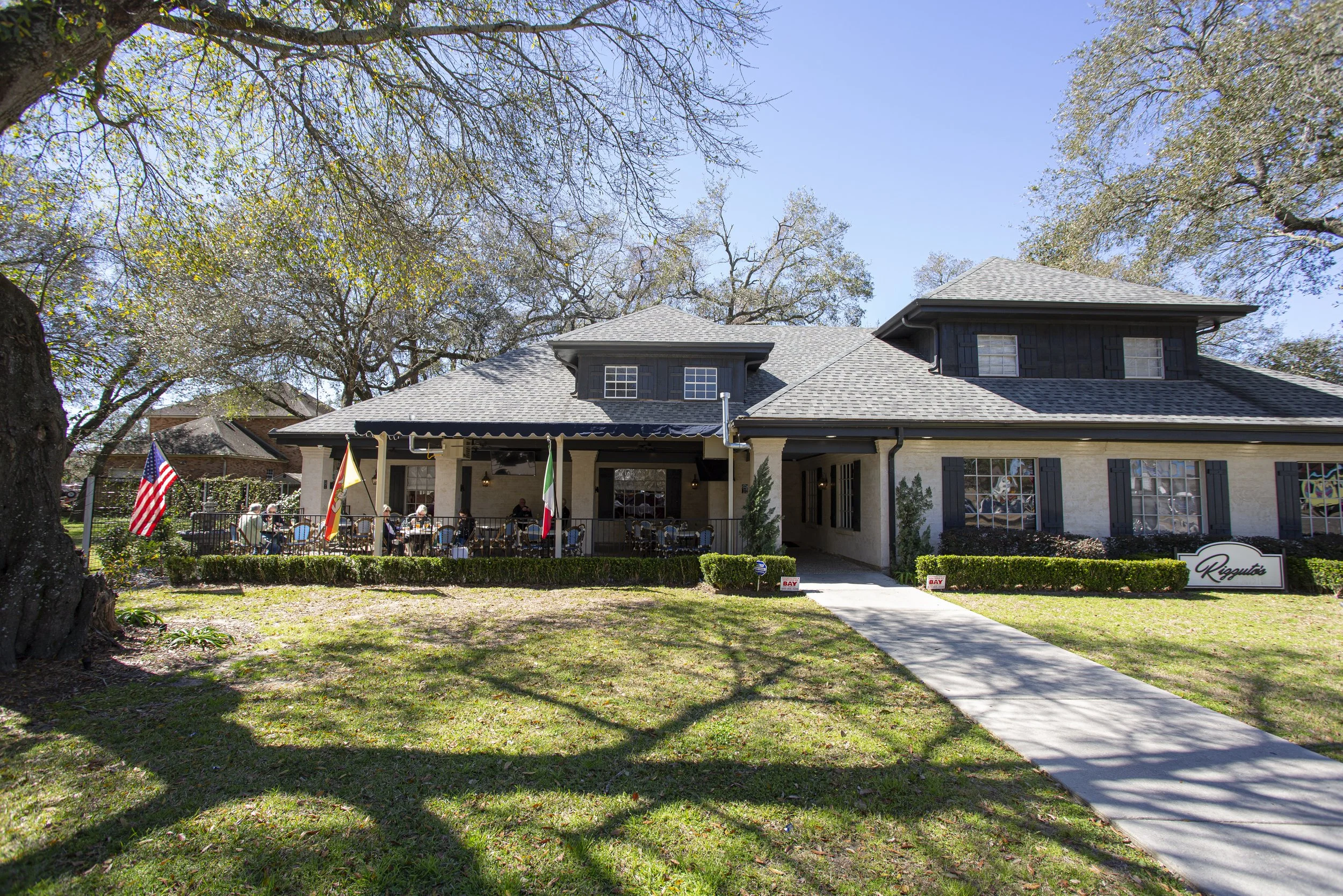 A house with a large porch, black shutters, and a gray roof. There are several people sitting outdoors on the porch and tables, and flags in front of the house. The yard has green grass and a sidewalk leading to the house. Tall trees with some leaves in the background and a clear blue sky overhead.