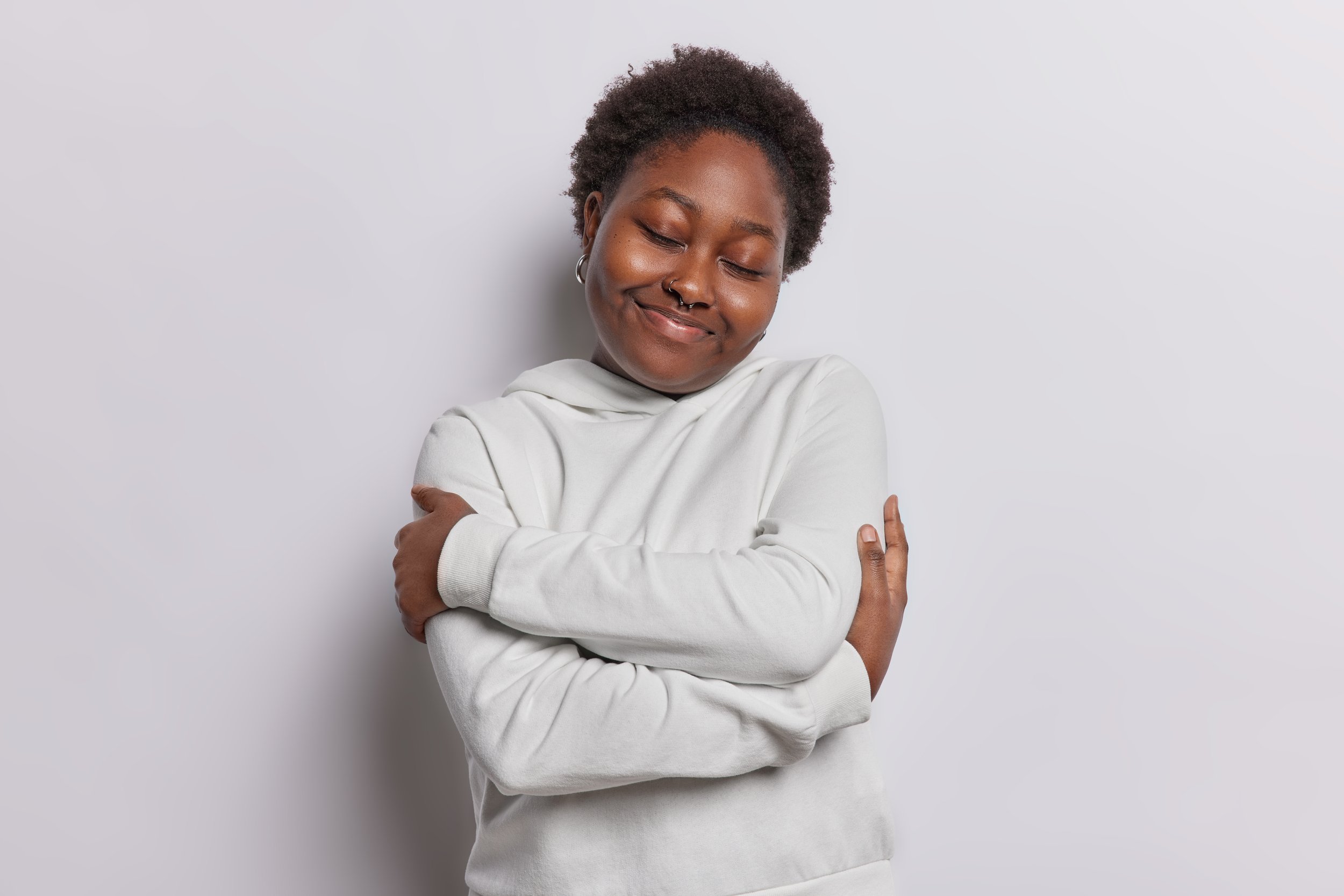 Young woman with short curly hair and nose piercings smiling with eyes closed, hugging herself against a plain light gray background.