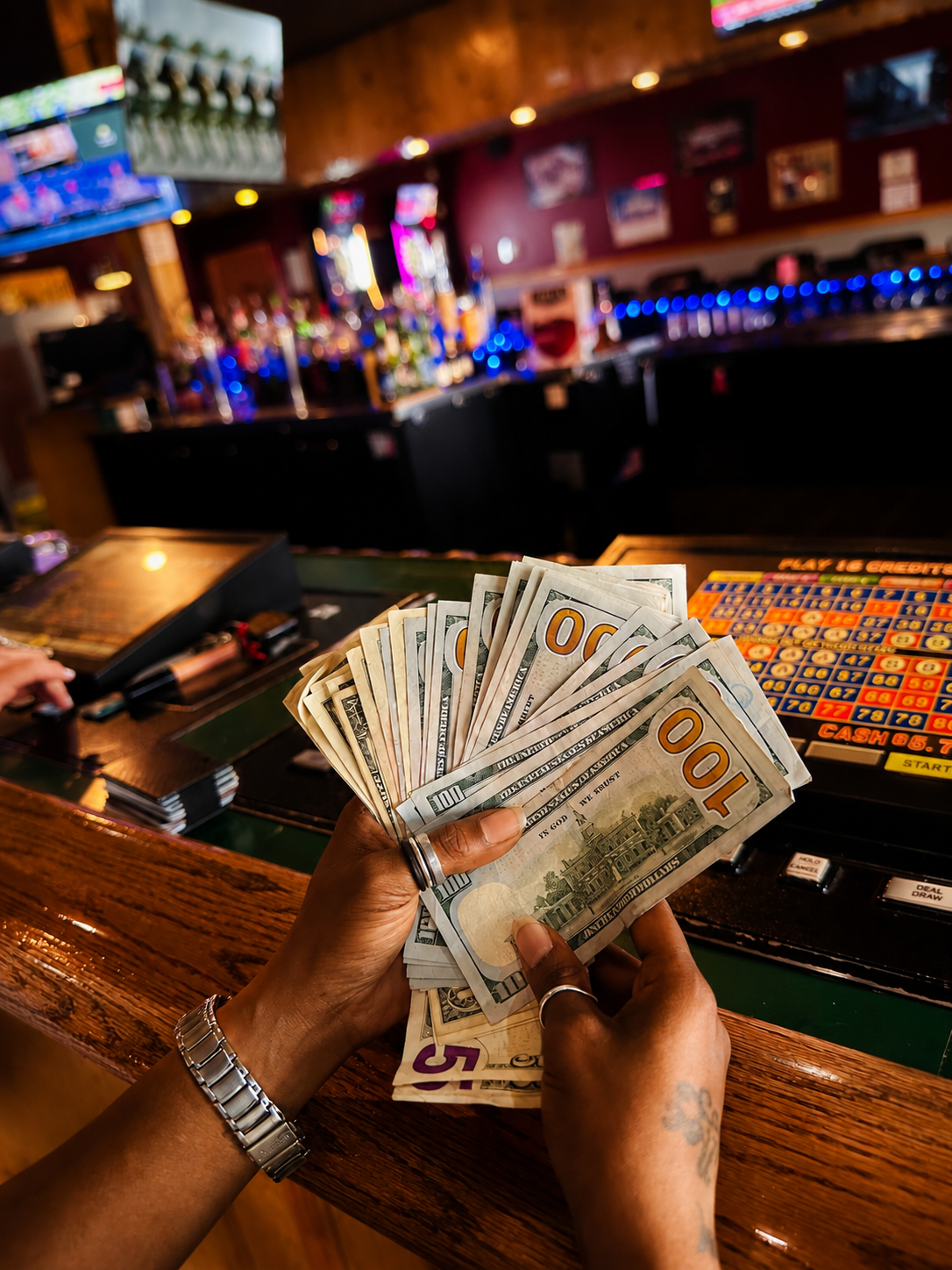 Person holding a fan of cash at a bar with a gambling machine in the background.