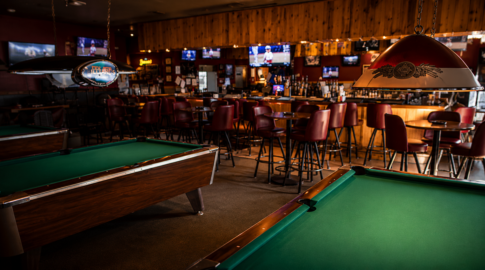 Interior of a bar or pub with pool tables, bar stools, high tables, and multiple TV screens on the walls, dim lighting, and a wooden decor.
