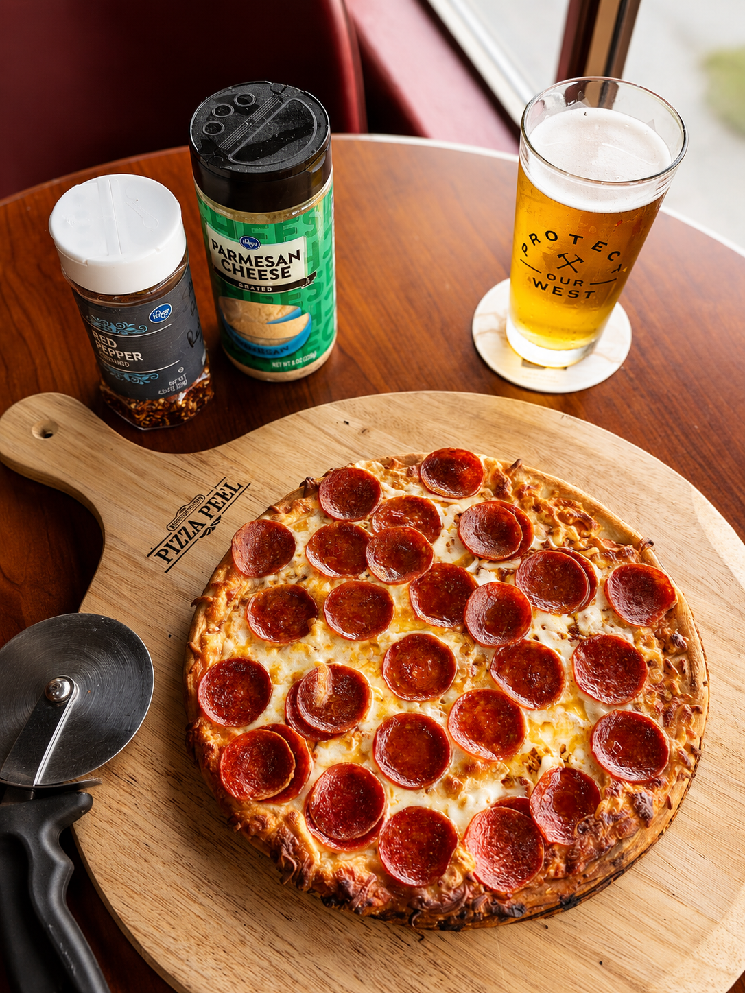 Pepperoni pizza on a wooden pizza peel, alongside a glass of beer, and shakers of parmesan cheese and red pepper flakes on a wooden table.