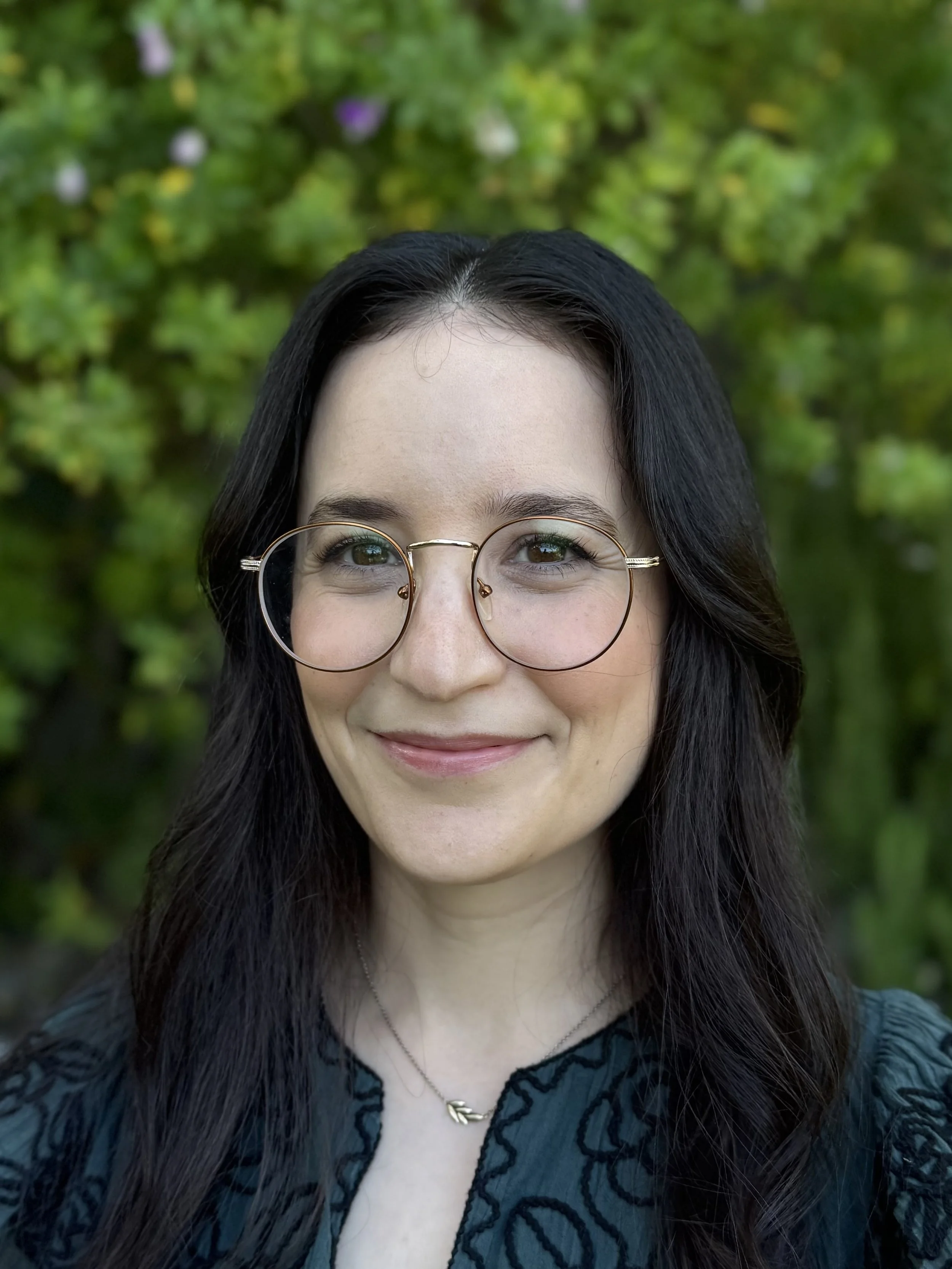 A person with long black hair and glasses smiling outdoors, with green foliage in the background.