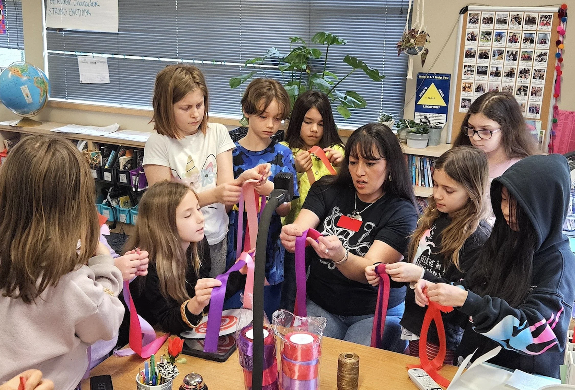 Photograph of a female teacher with long dark hair demonstrating ribbon tying with a mixed gender group of eight students of late elementary age