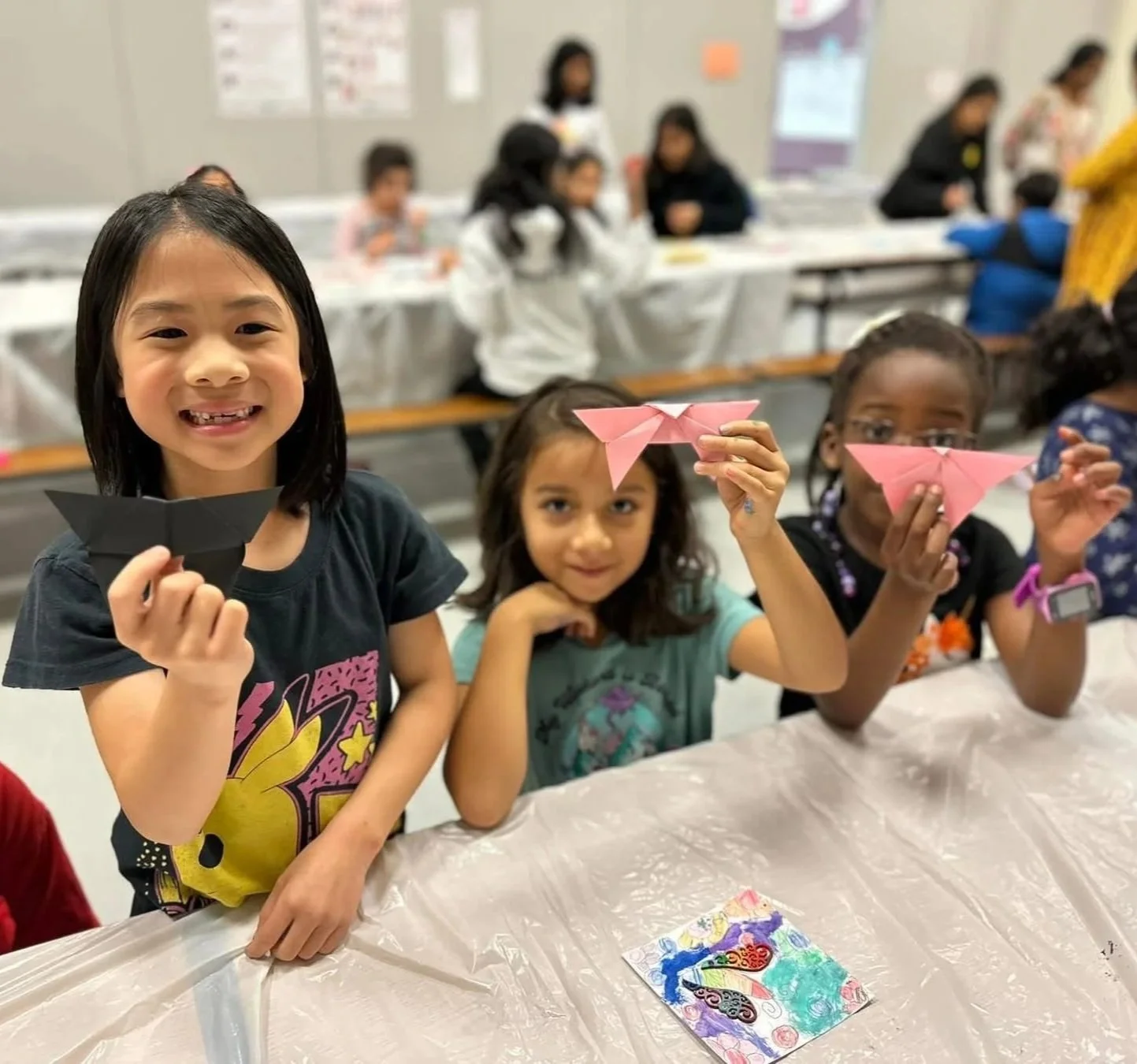 Photograph of three elementary age girls showing origami creations to the camera and smiling
