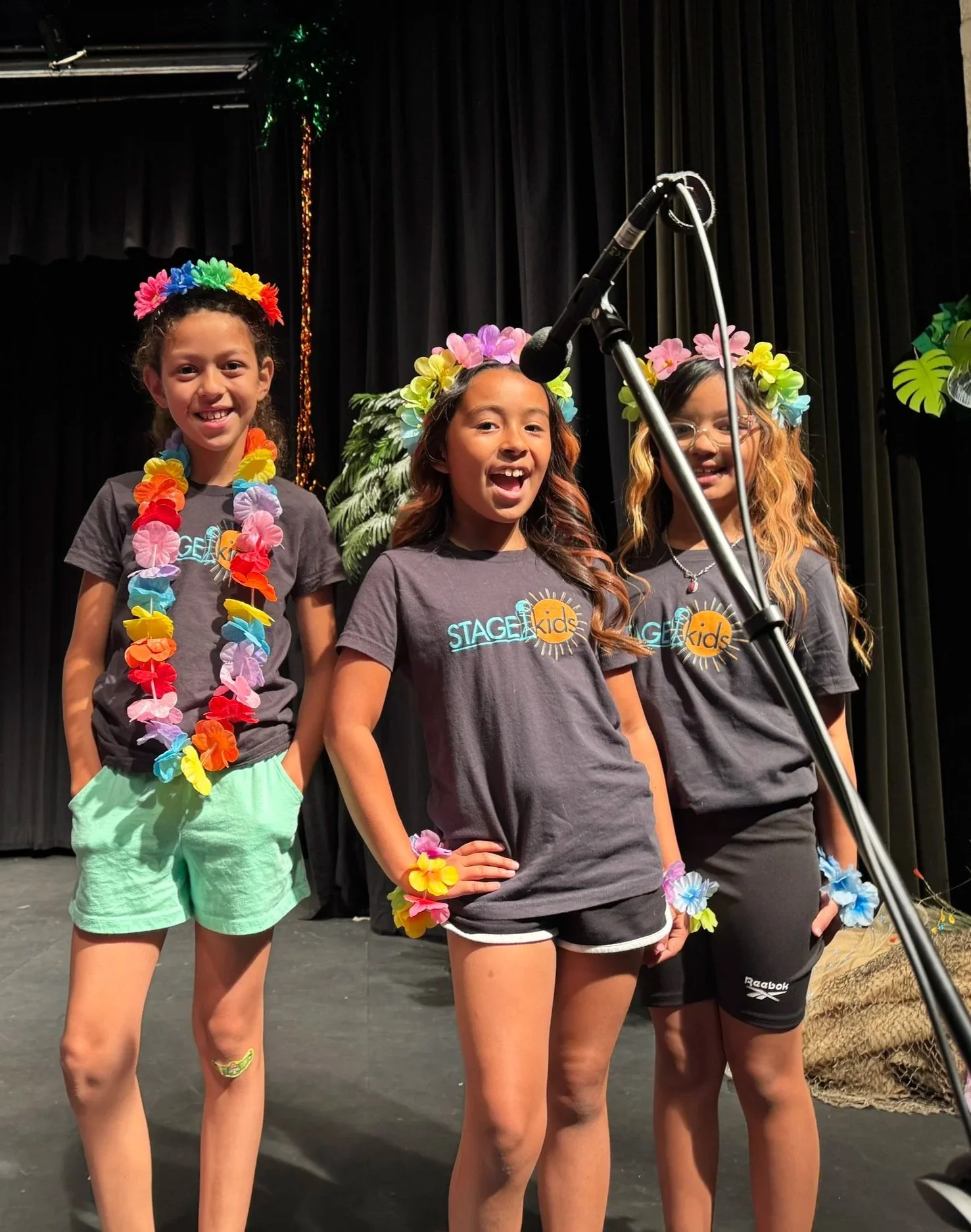 Photograph of three elementary age girls singing into a shared microphone and wearing plastic leis