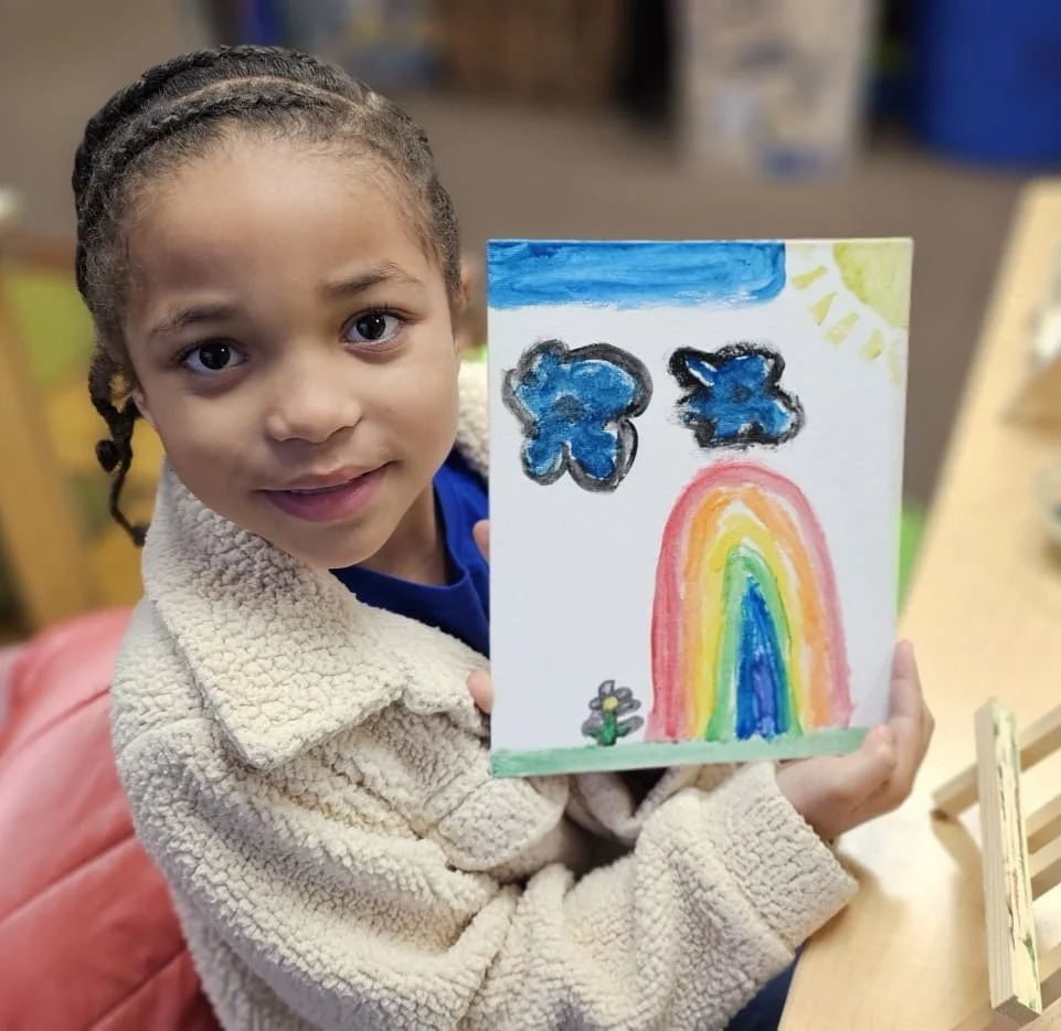 Photograph of a young elementary age girl with braids showing a painting of a rainbow, clouds and sun to the camera and smiling