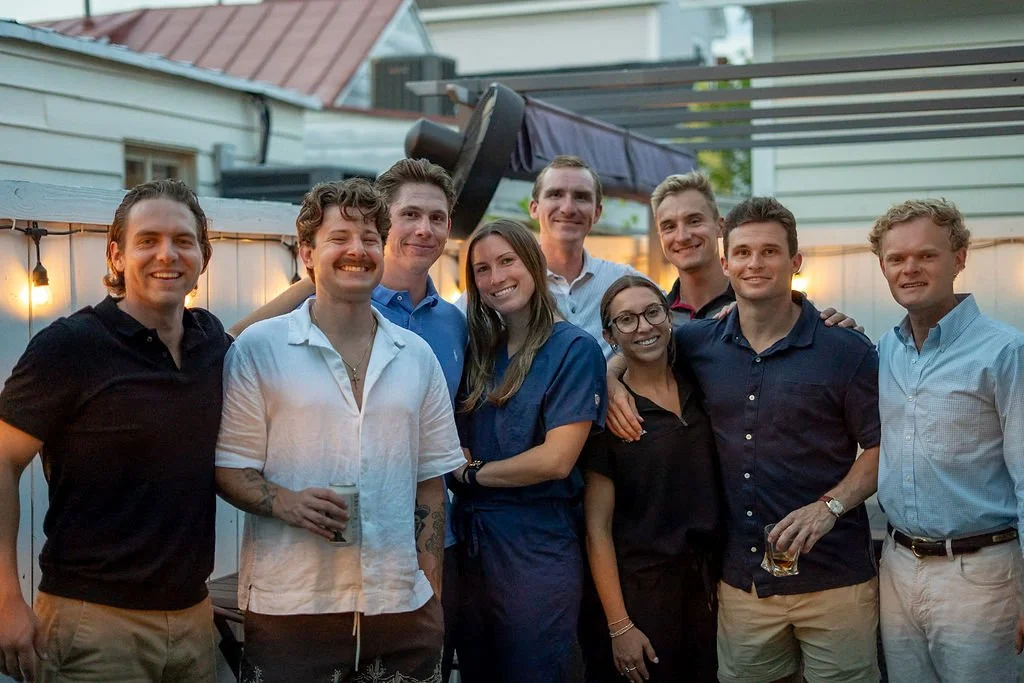 A group of eight young adults gathered closely outdoors during sunset, smiling and posing for a photo, with string lights and a wooden fence in the background.