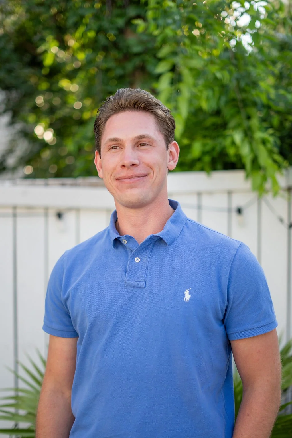 A young man with light skin and brown hair smiling outdoors, wearing a blue polo shirt with a small white logo, standing in front of green foliage and a white fence.
