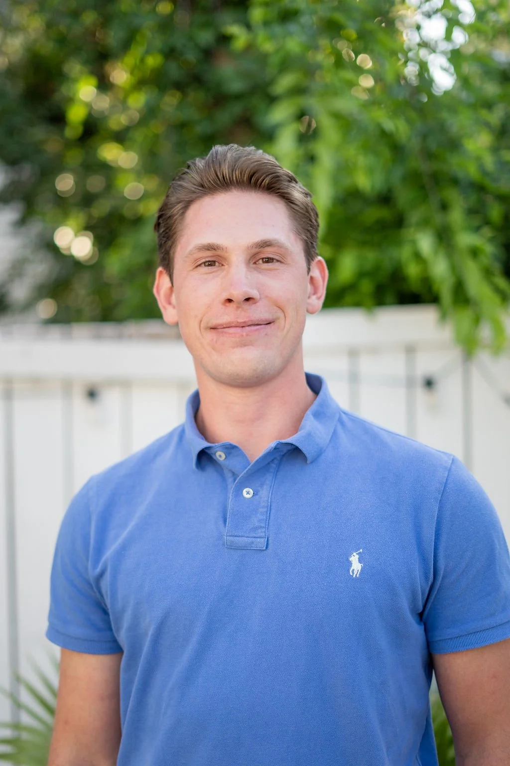 A young man with light skin, brown hair, and a slight smile, wearing a blue polo shirt, standing outdoors with green trees and a white fence in the background.