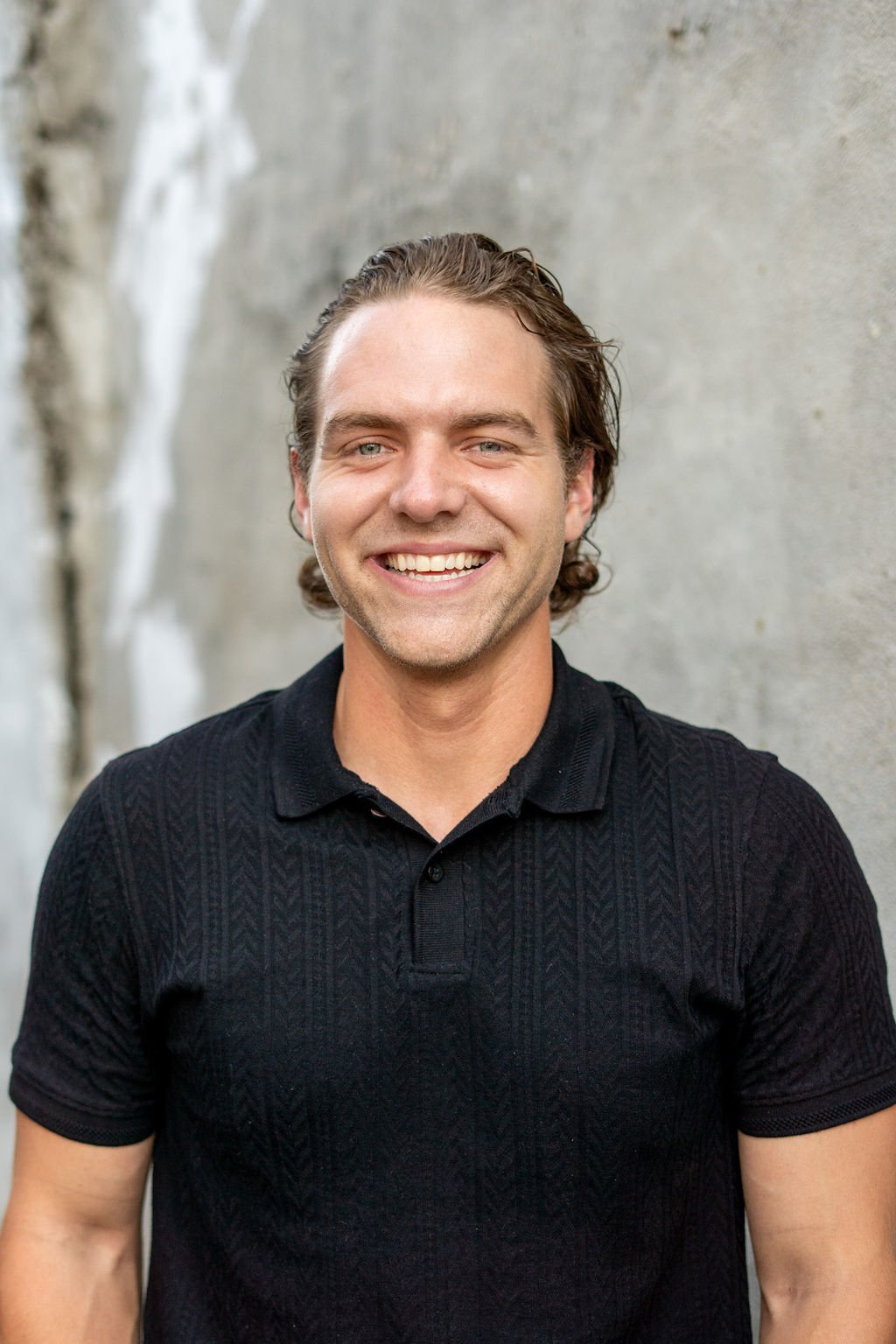 A young man with wavy brown hair and a wide smile, wearing a black short-sleeved polo shirt, standing outdoors against a blurred light-colored wall.