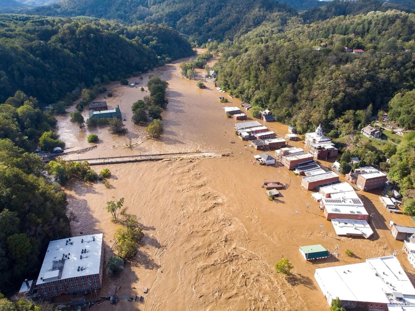 Aerial view of a small town surrounded by green hills, with a river overflowing and flooding the streets and buildings.