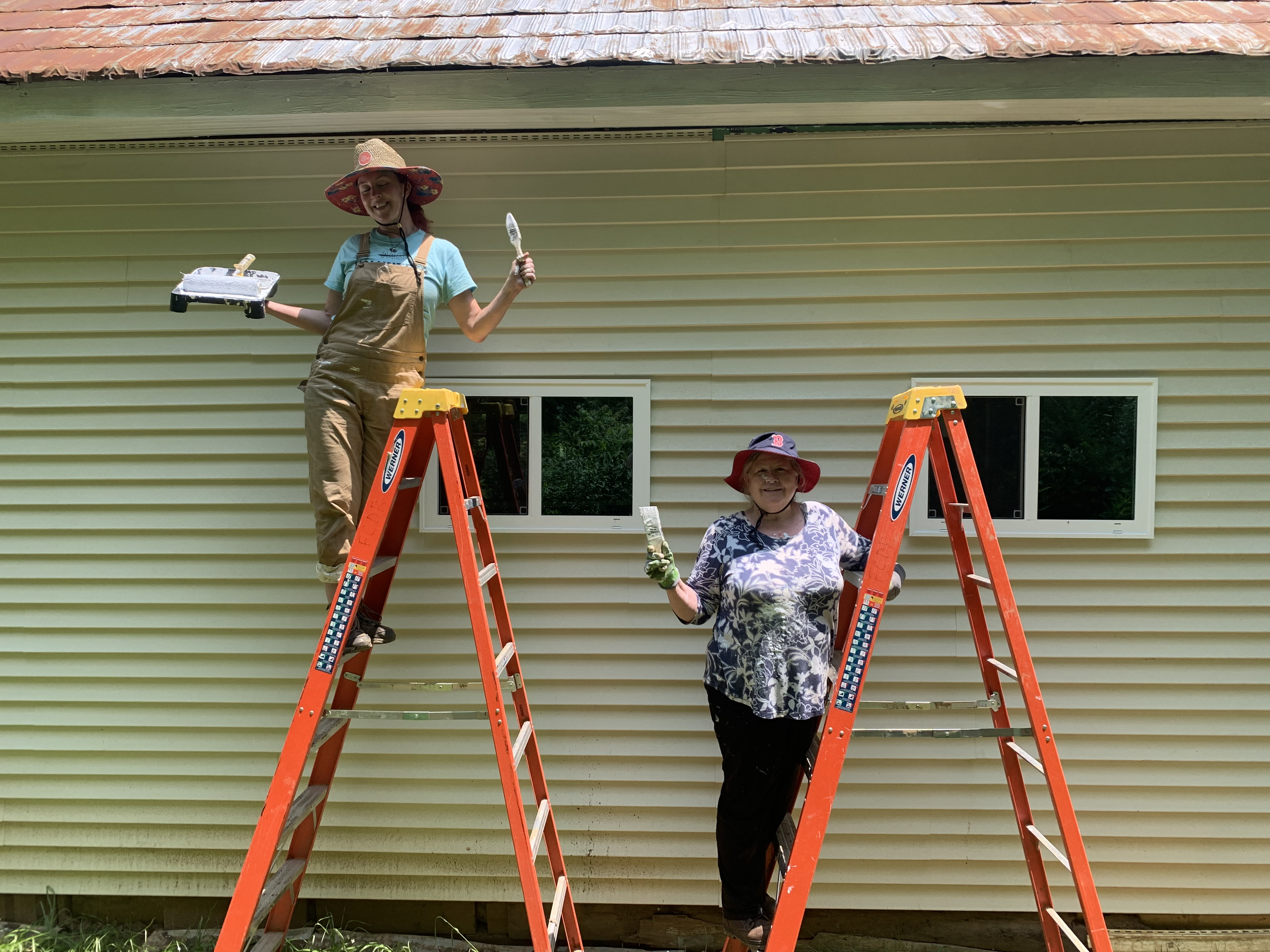 Two women on orange ladders painting the exterior of a house with yellow siding. One woman stands on a ladder holding a paintbrush and a paint tray, wearing tan overalls and a wide-brimmed straw hat. The other woman stands on a ladder wearing a patterned blouse and a red bucket hat, holding a paintbrush.
