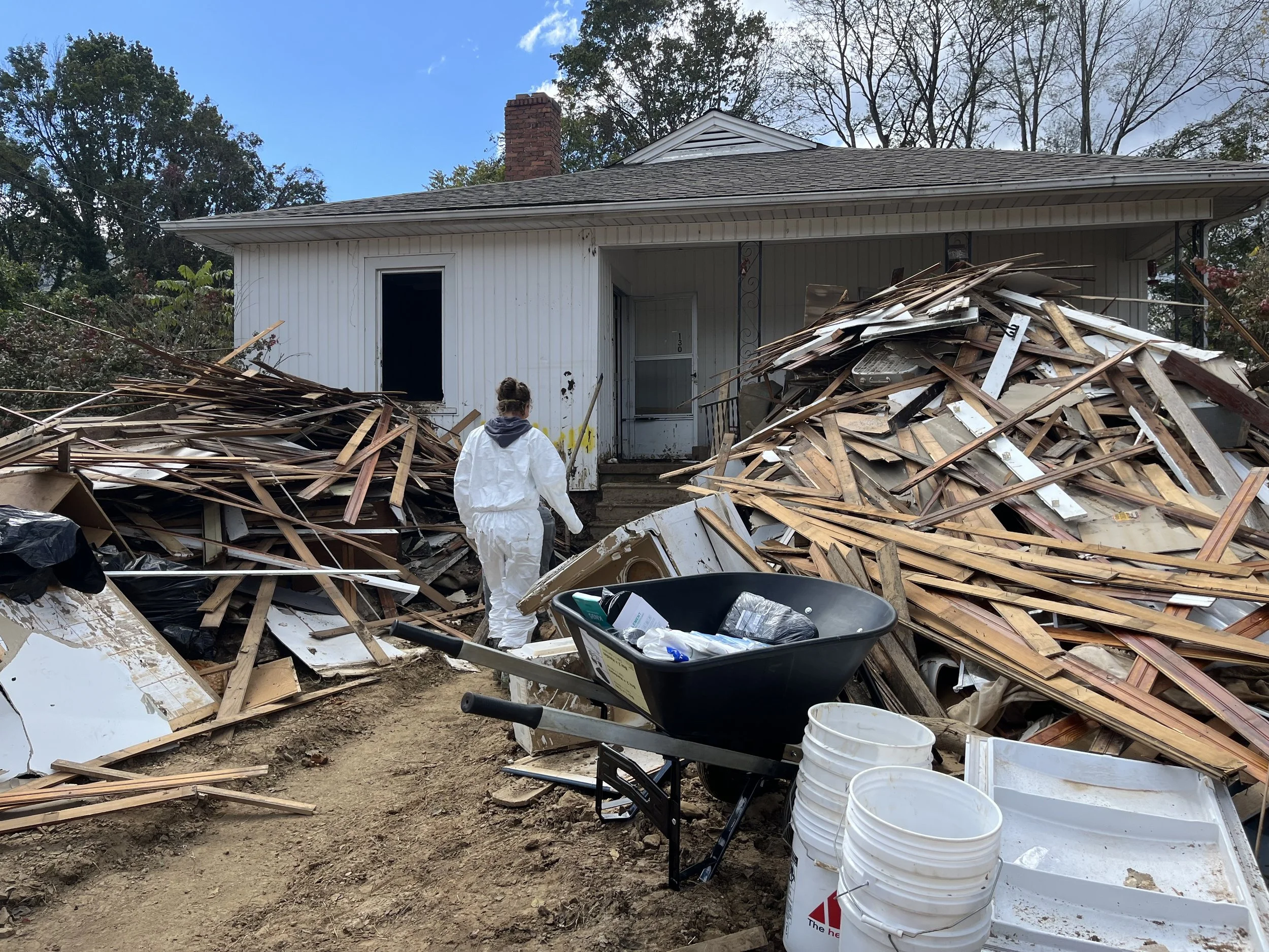 A person wearing white protective clothing stands in front of a heavily damaged house with debris, wood, and wreckage scattered on the ground. There are buckets, a wheelbarrow, and other tools nearby.