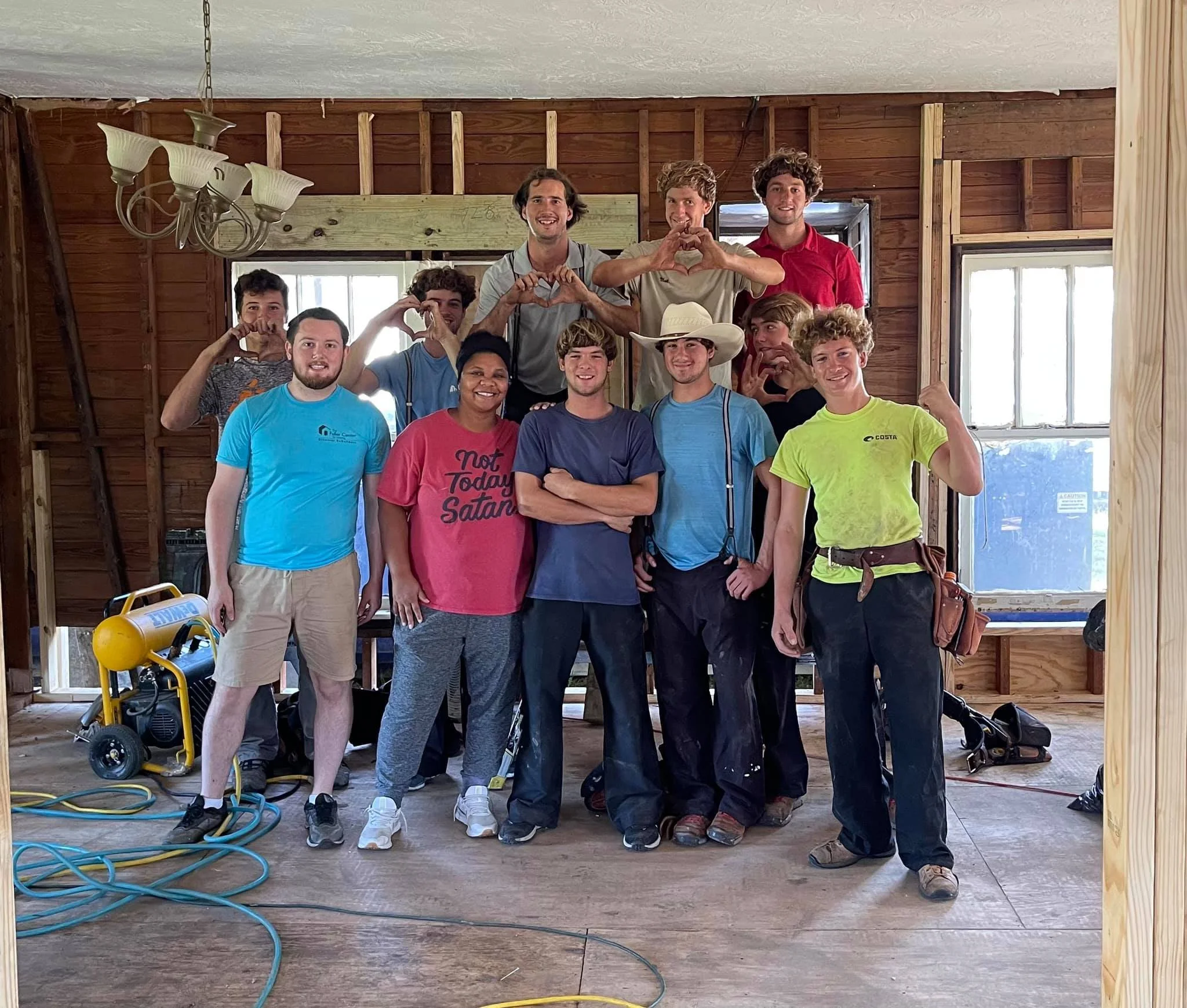 A group of young people, mostly men, standing inside a partially constructed wooden building. They are smiling, some making hand hearts, and are dressed casually. Construction tools and wires are visible on the floor.