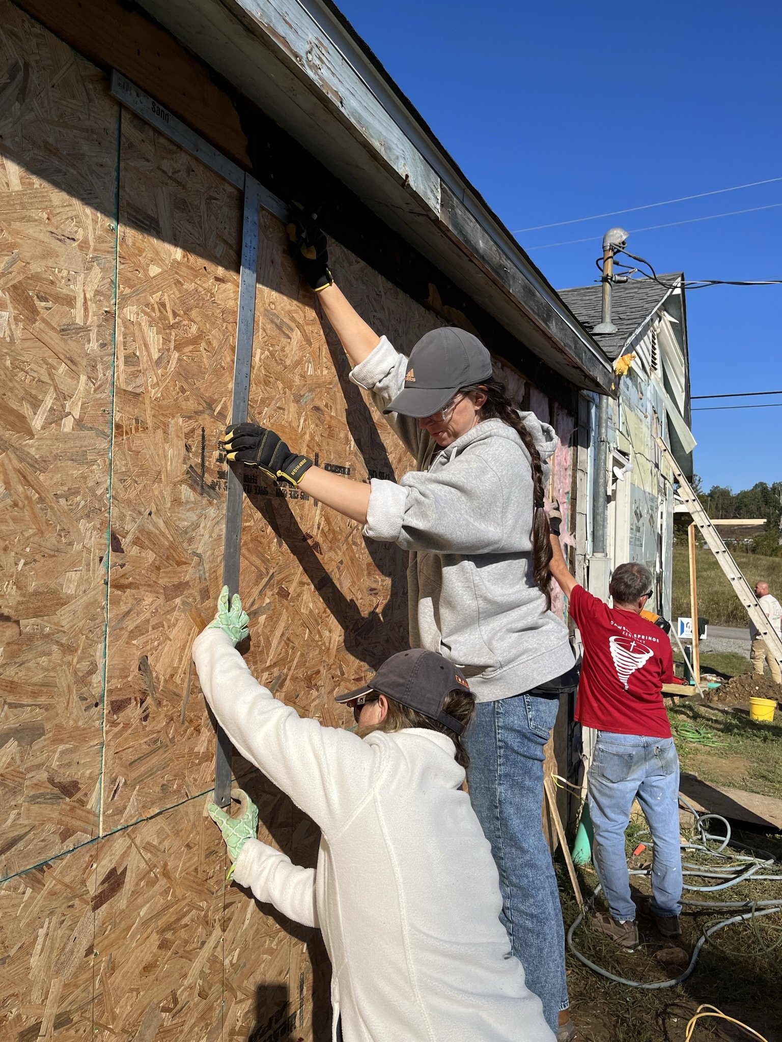 A group of people are working together to install or repair the exterior of a building. They are attaching insulation or sheathing to the wall. The scene takes place outdoors on a bright, sunny day.