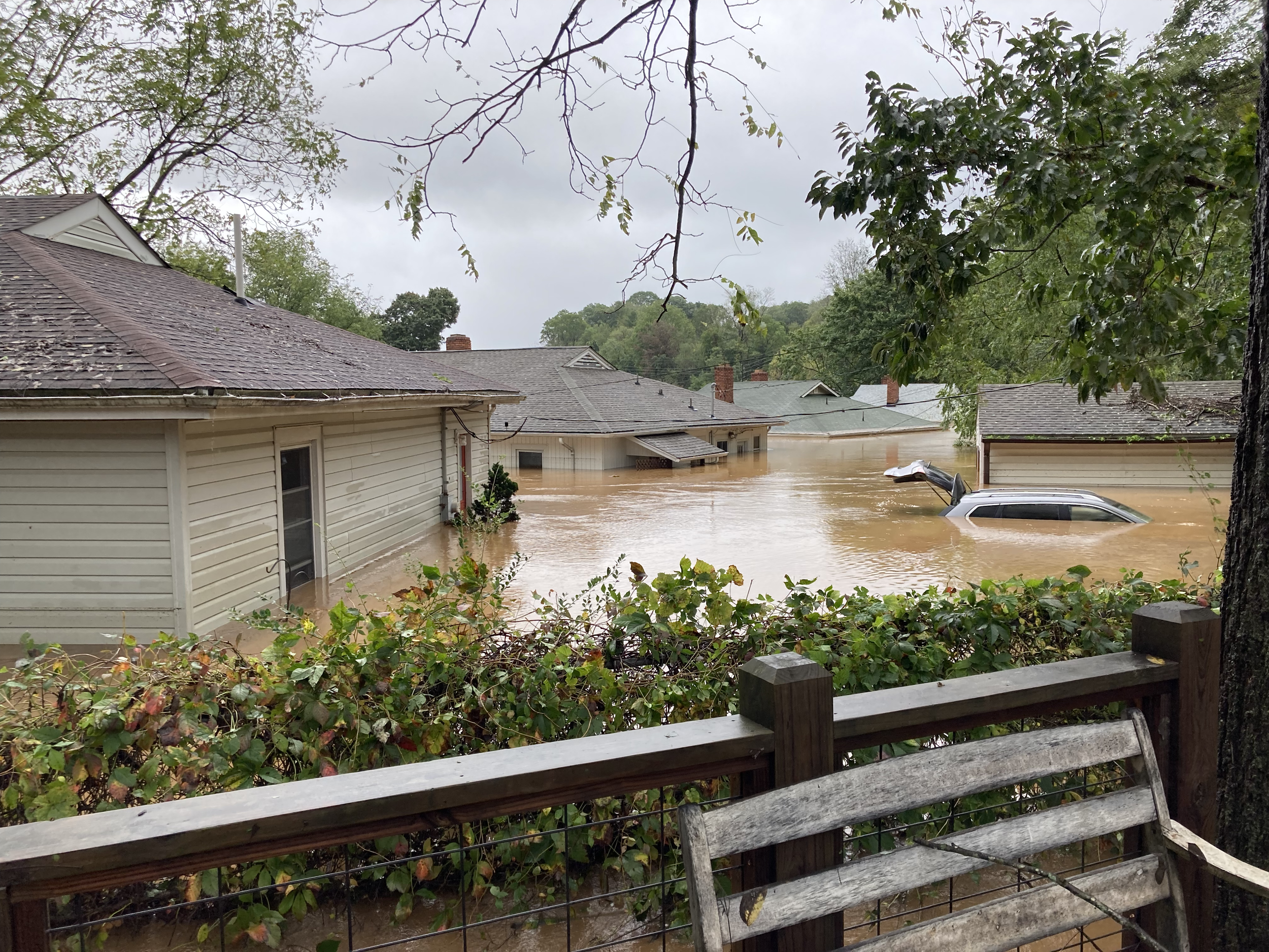 Flooded neighborhood with partially submerged vehicles, houses, trees, and overcast sky after heavy rain.