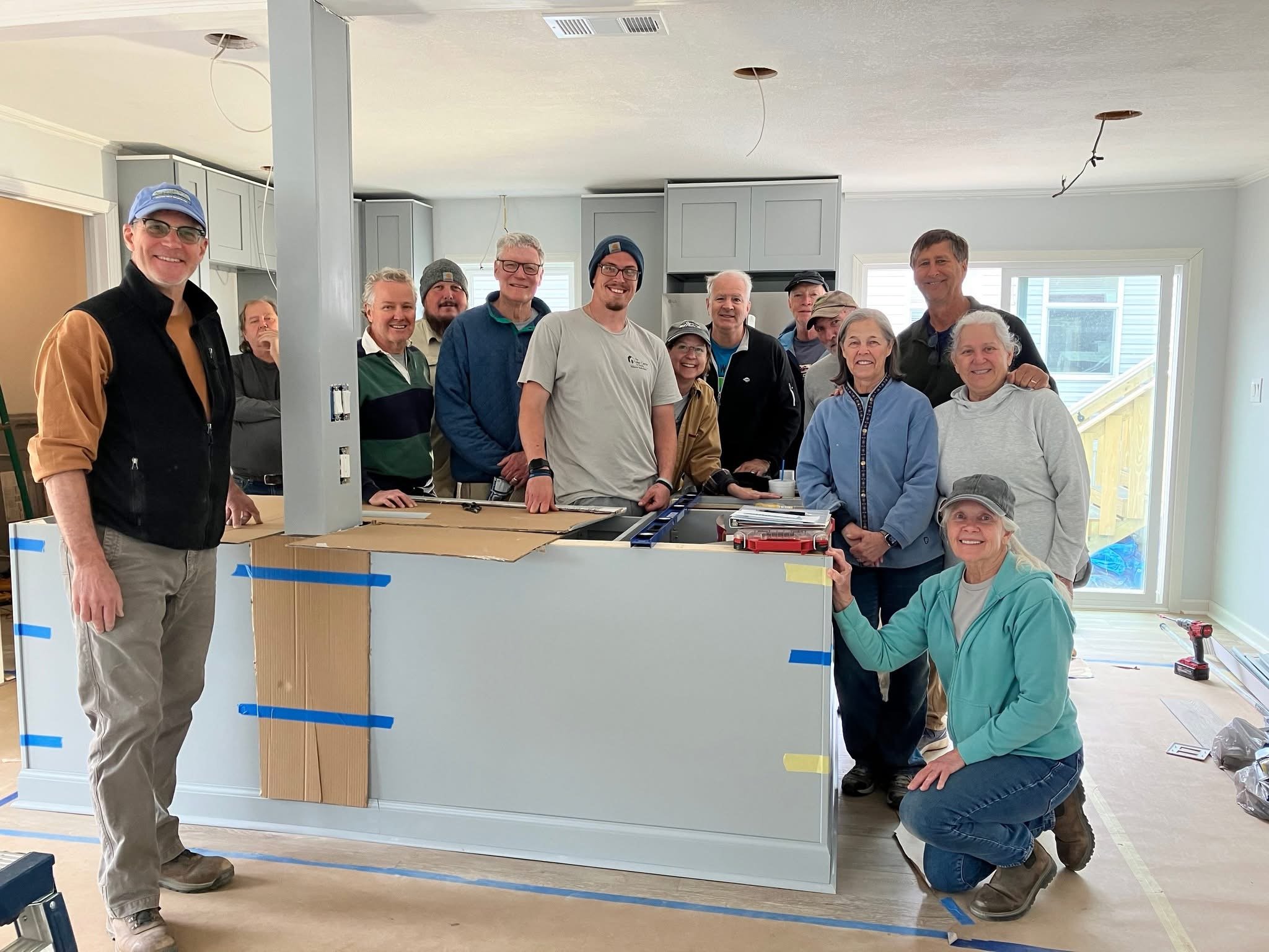 Group of about 16 people gathered indoors around a partially built wooden structure, smiling for the camera during a construction or renovation project.