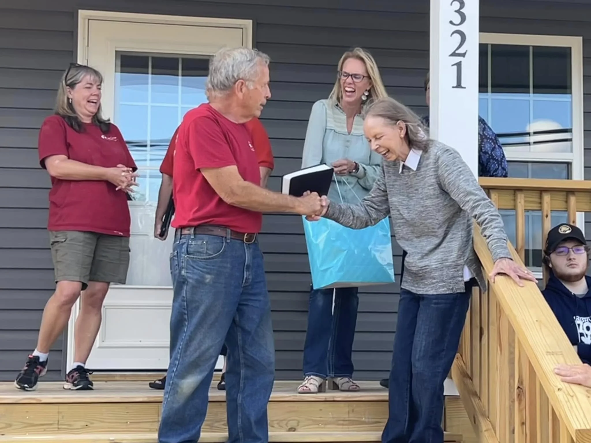 People gathered on a porch, with a man and a woman shaking hands and laughing, while others observe and chat, in front of a house with dark siding and white trim.