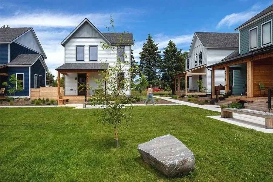 A residential neighborhood with modern houses, a grassy lawn, a young tree, and a large rock in the foreground. The houses are painted blue, white, and teal, with wooden porches and steps. A person is walking along the sidewalk.
