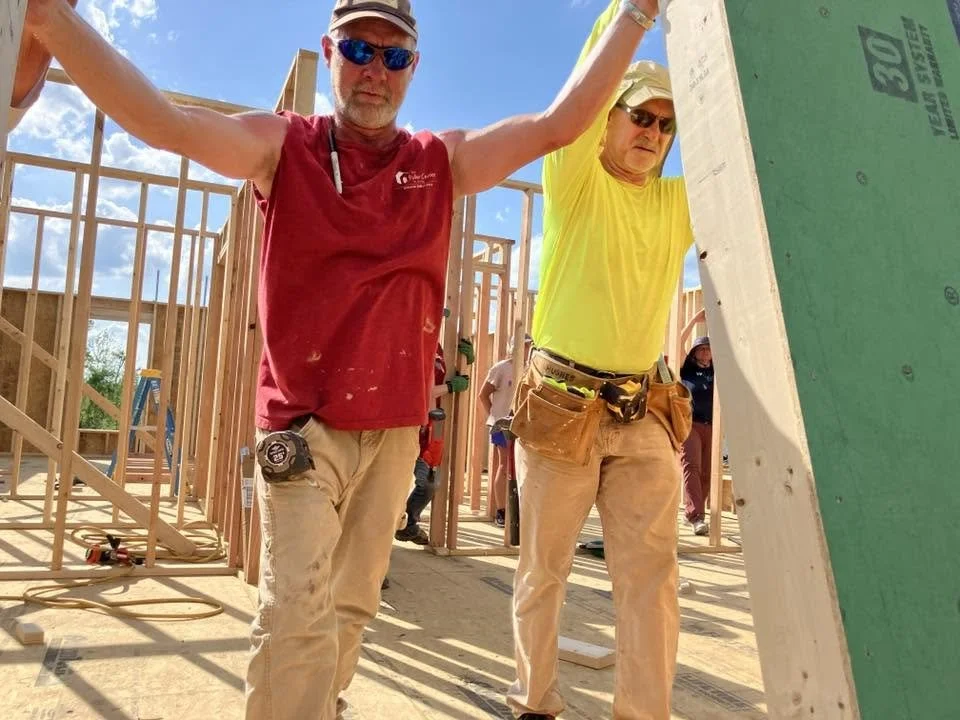 Two construction workers on a building site, framing wooden walls under a blue sky with some clouds. One worker, wearing a red sleeveless shirt, sunglasses, and a cap, is standing with arms raised. The other, wearing a yellow shirt and sunglasses, is leaning over a large green board.