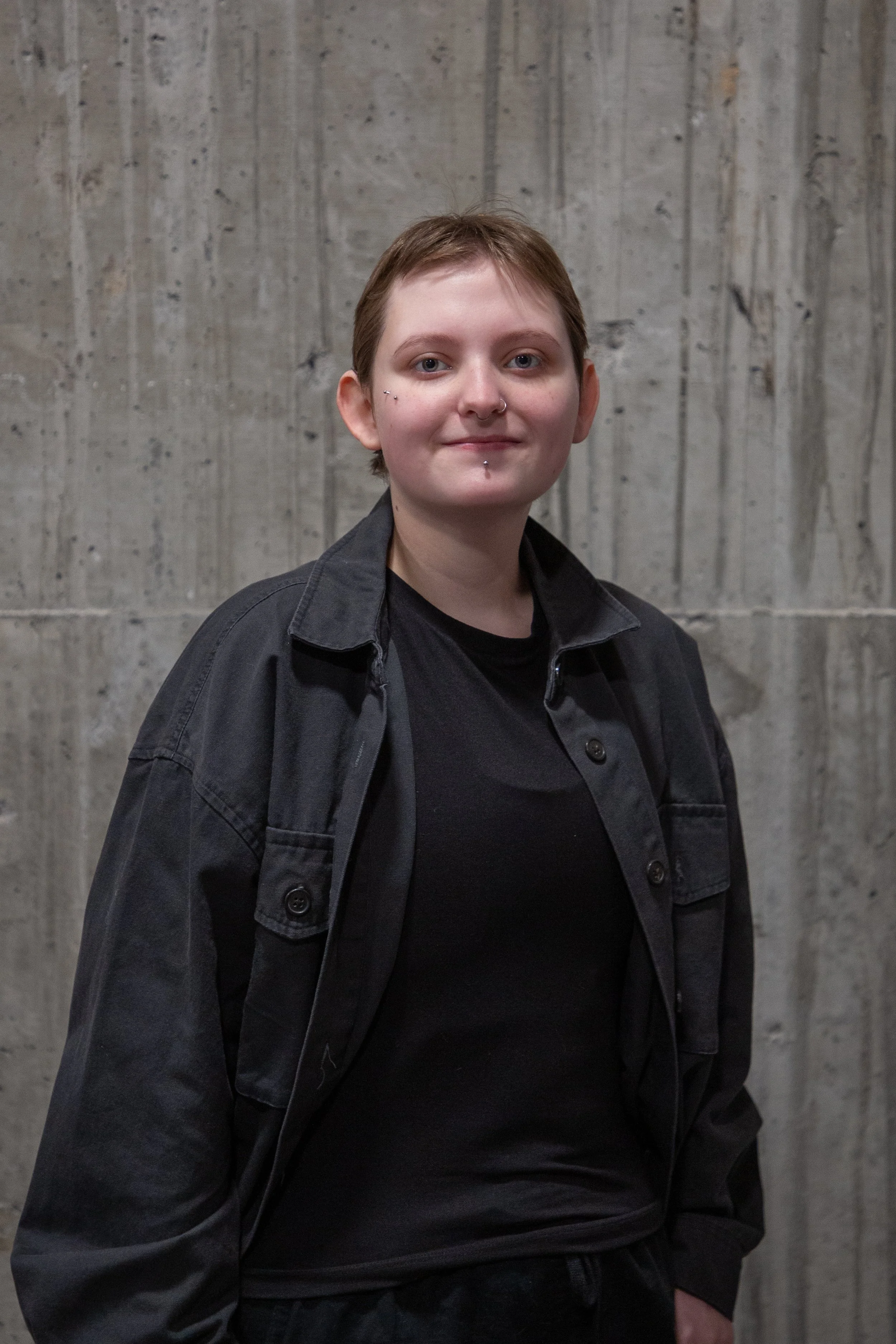 A person with short brown hair, multiple facial piercings, and light skin, wearing a black shirt and black jacket, standing against a textured concrete wall.