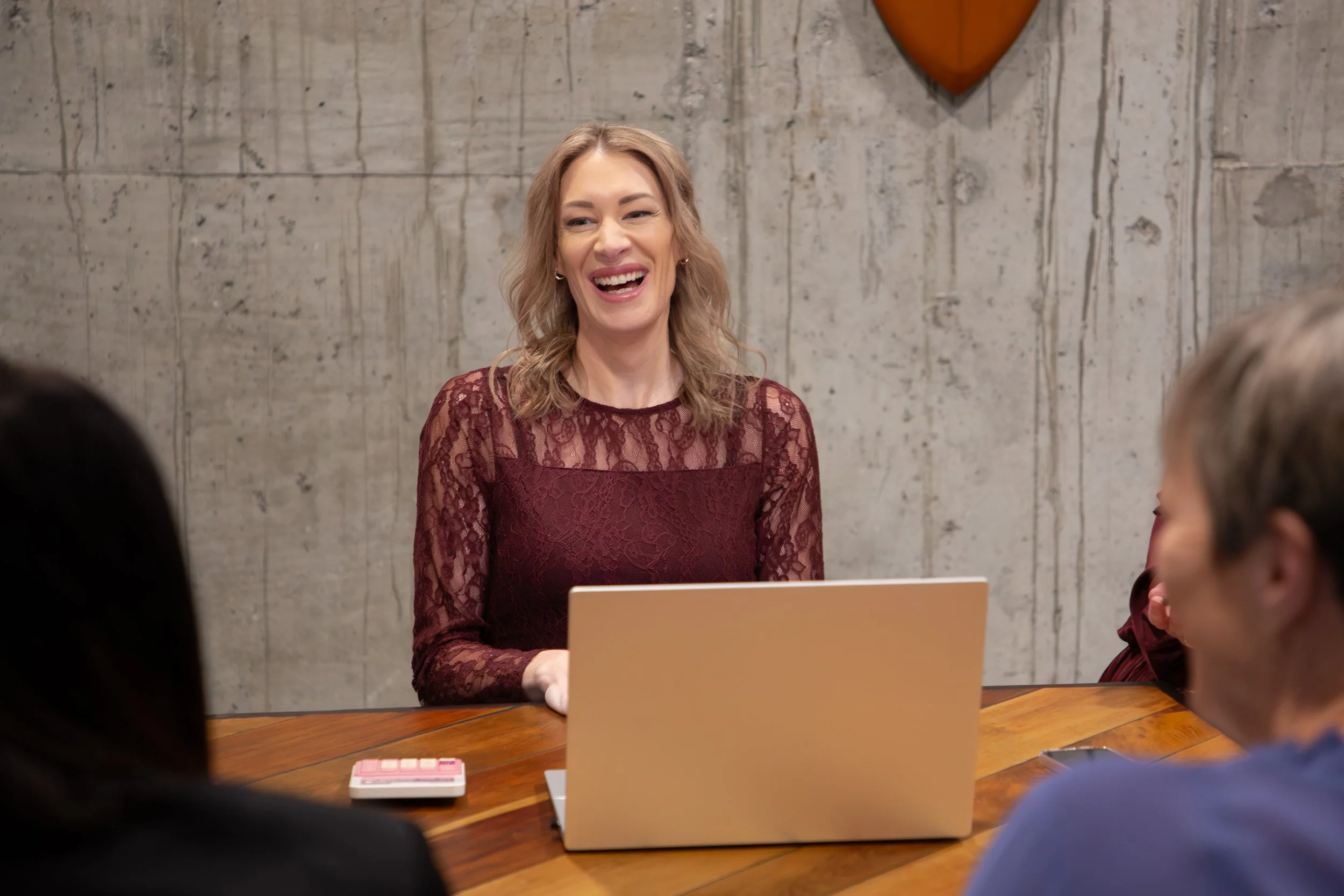 A woman in a maroon lace dress sitting at a wooden table with a laptop, smiling and talking to people in a room with a concrete wall and a heart-shaped decoration hanging on the wall.
