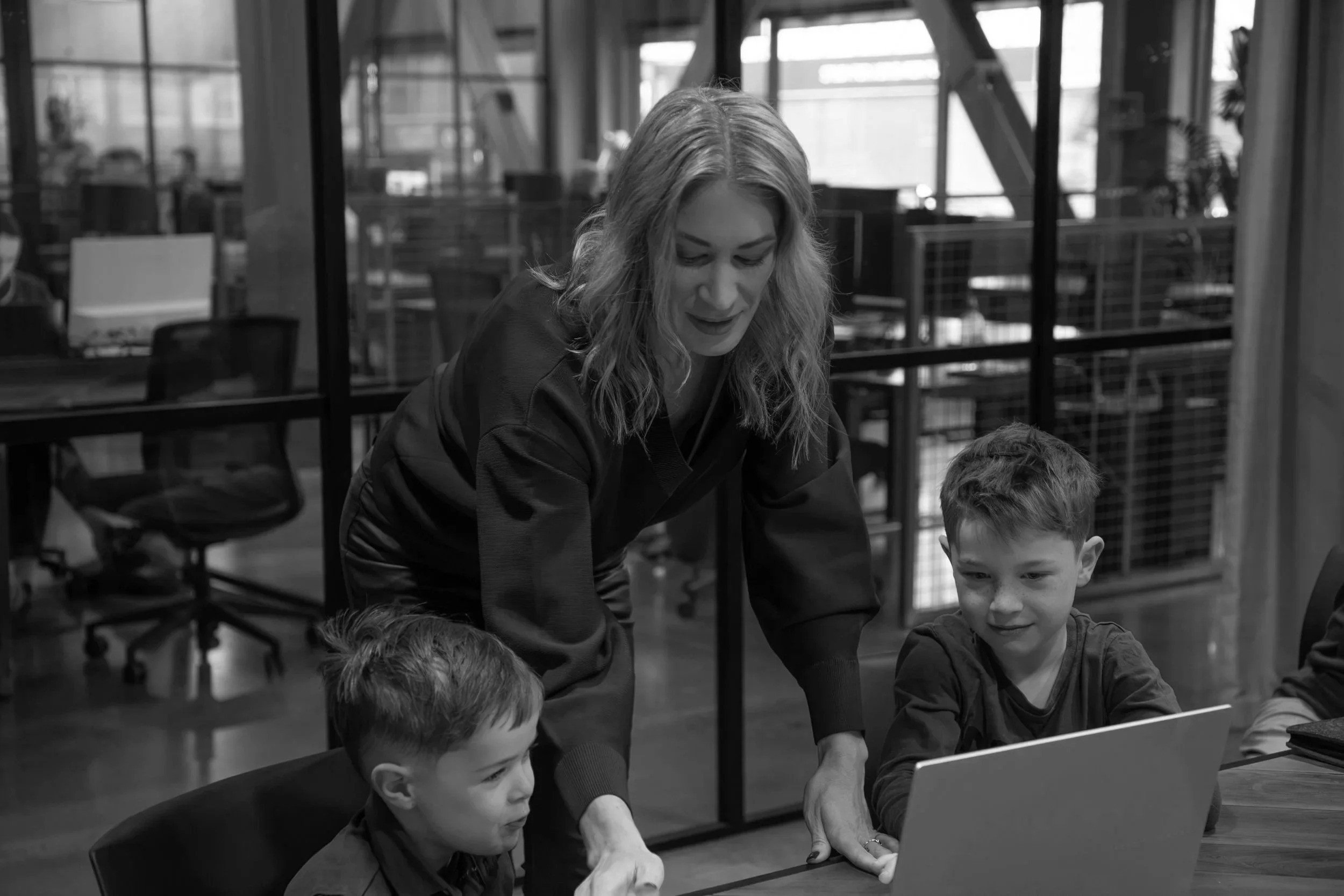 A woman with wavy hair leaning over a table, interacting with three young boys who are gathered around a laptop in a modern office setting.