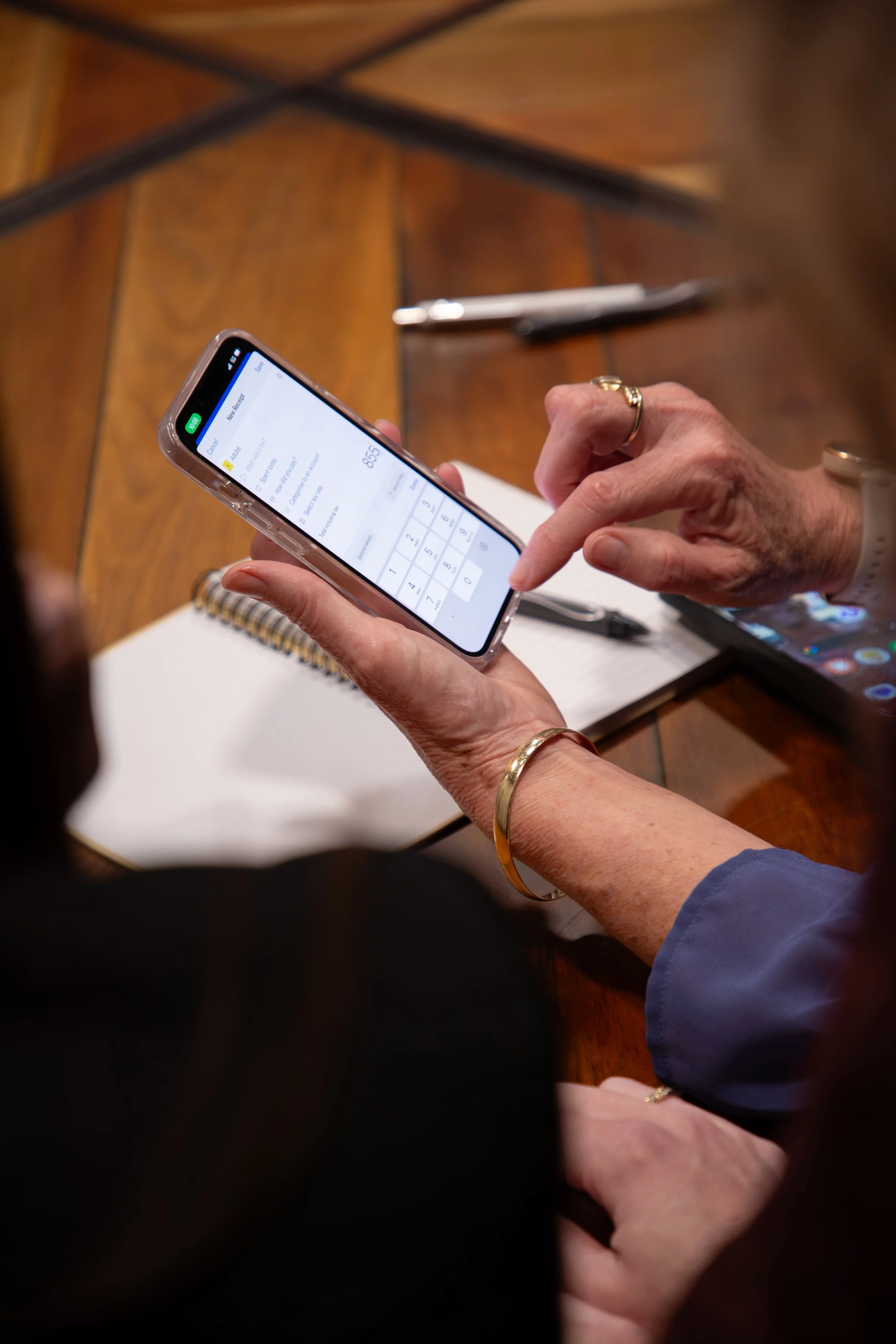 Close-up of a woman's hand with a gold bracelet and ring, holding a smartphone displaying a calculator app, on a wooden table with a notebook and pen.