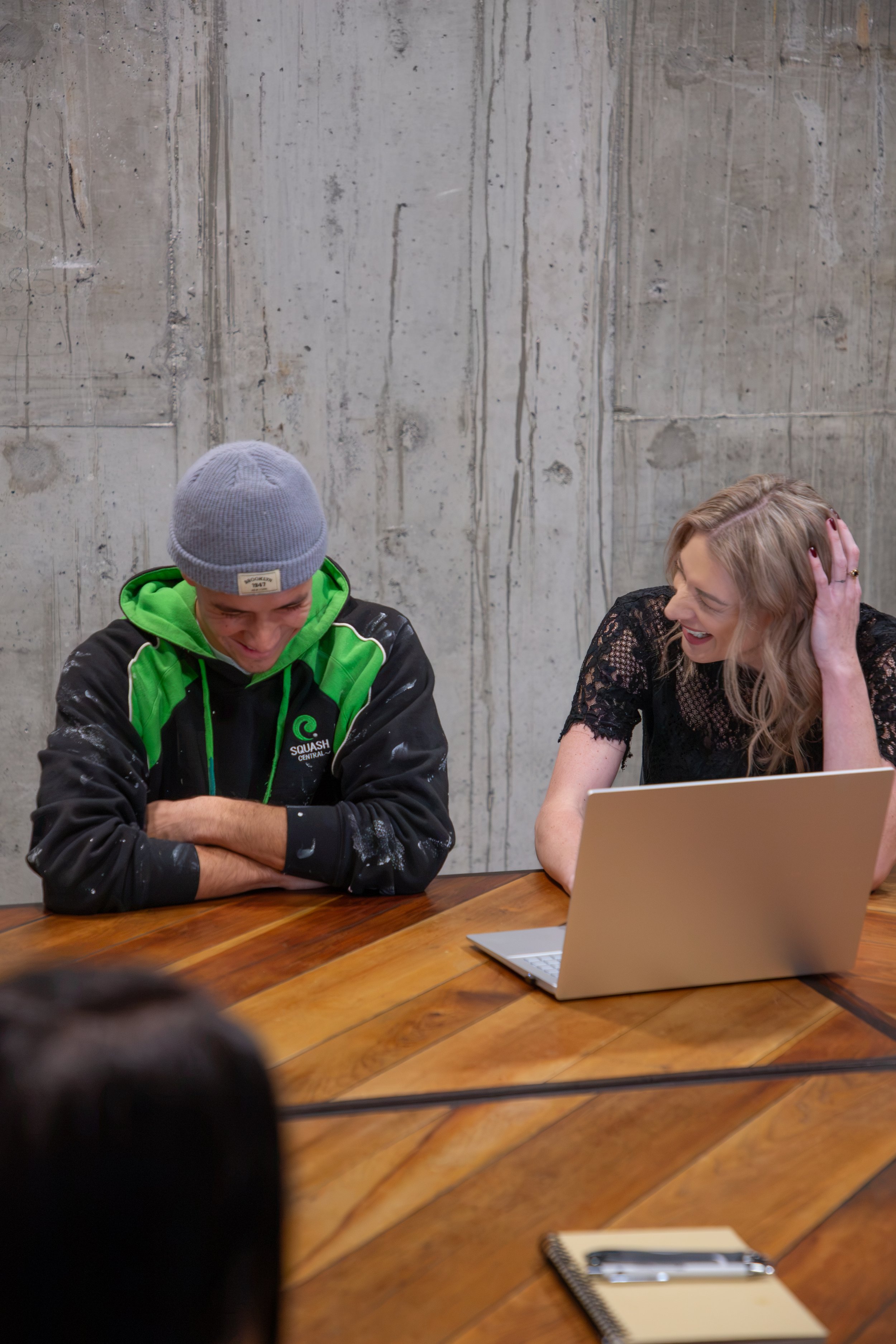 Two people sitting at a wooden table, both smiling and laughing. The person on the left is wearing a gray beanie and a black jacket with green accents, and the person on the right has blonde hair and is wearing a black lace top. There is a silver laptop and a spiral notebook with a pen on the table. The background features a concrete wall.