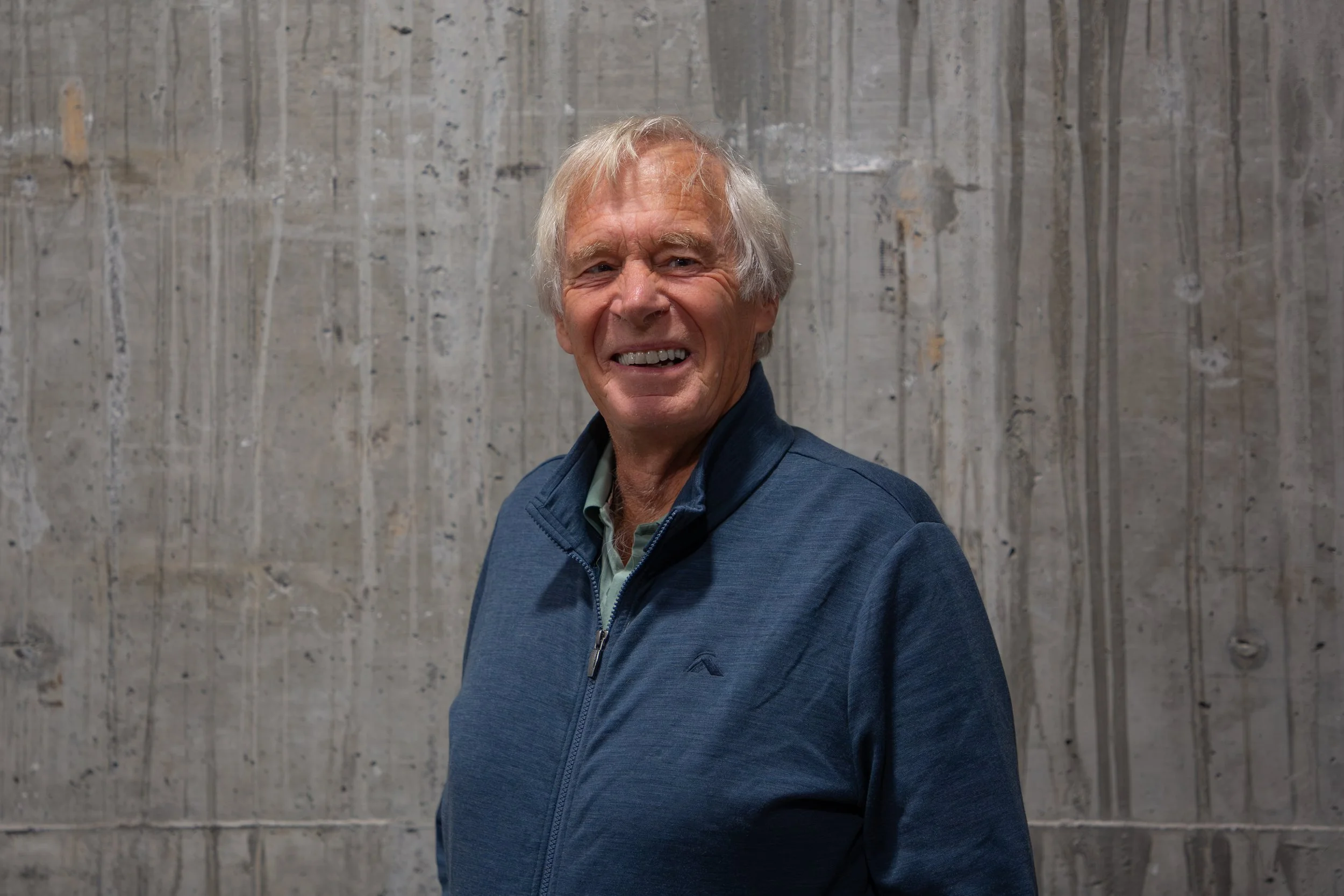 An elderly man with gray hair smiling and looking to the side, wearing a blue jacket, standing in front of a gray textured wall.