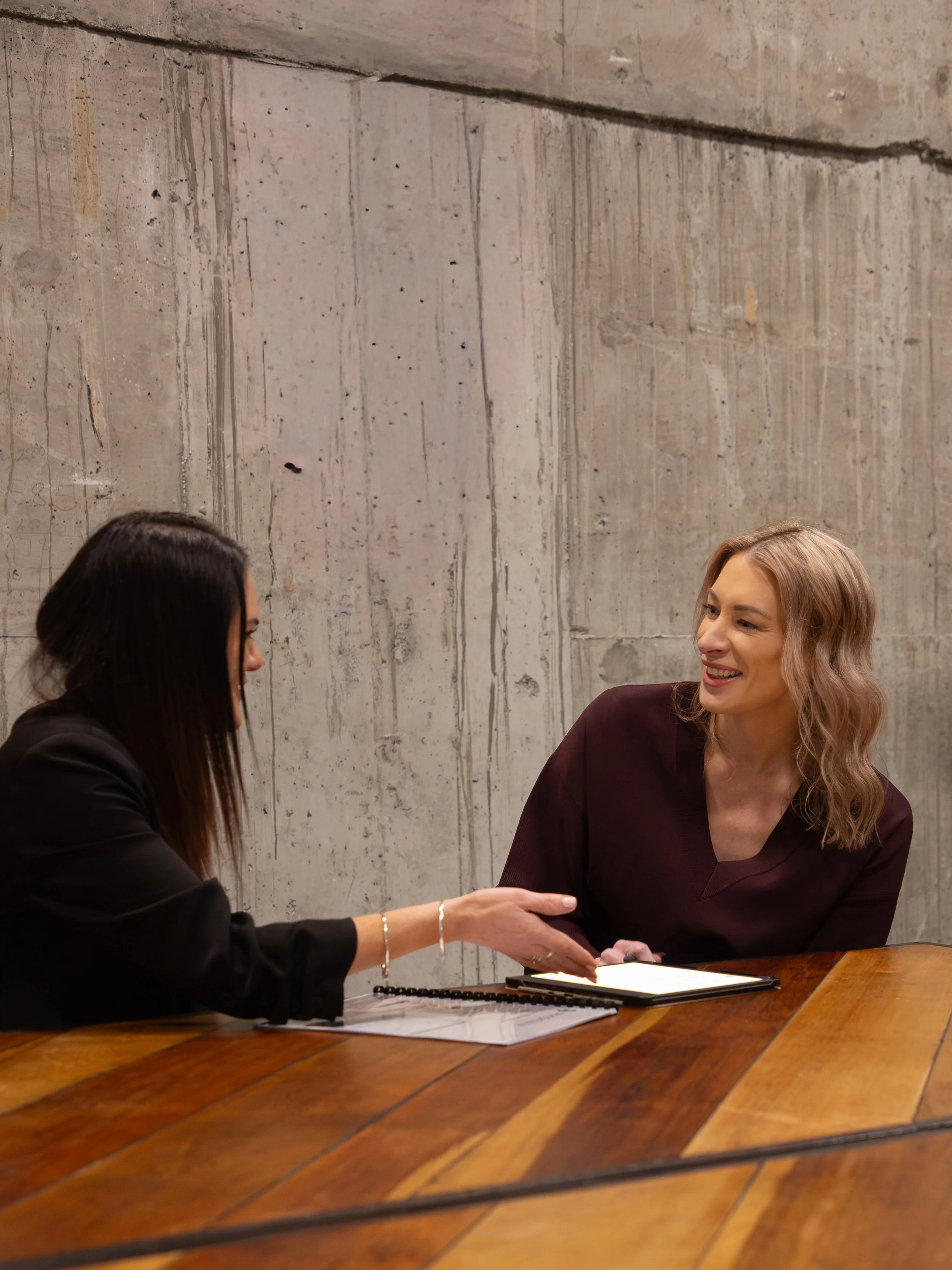Two women sitting at a wooden table, one with dark hair and glasses, the other with blonde hair, engaged in a conversation, with a gray concrete wall in the background.