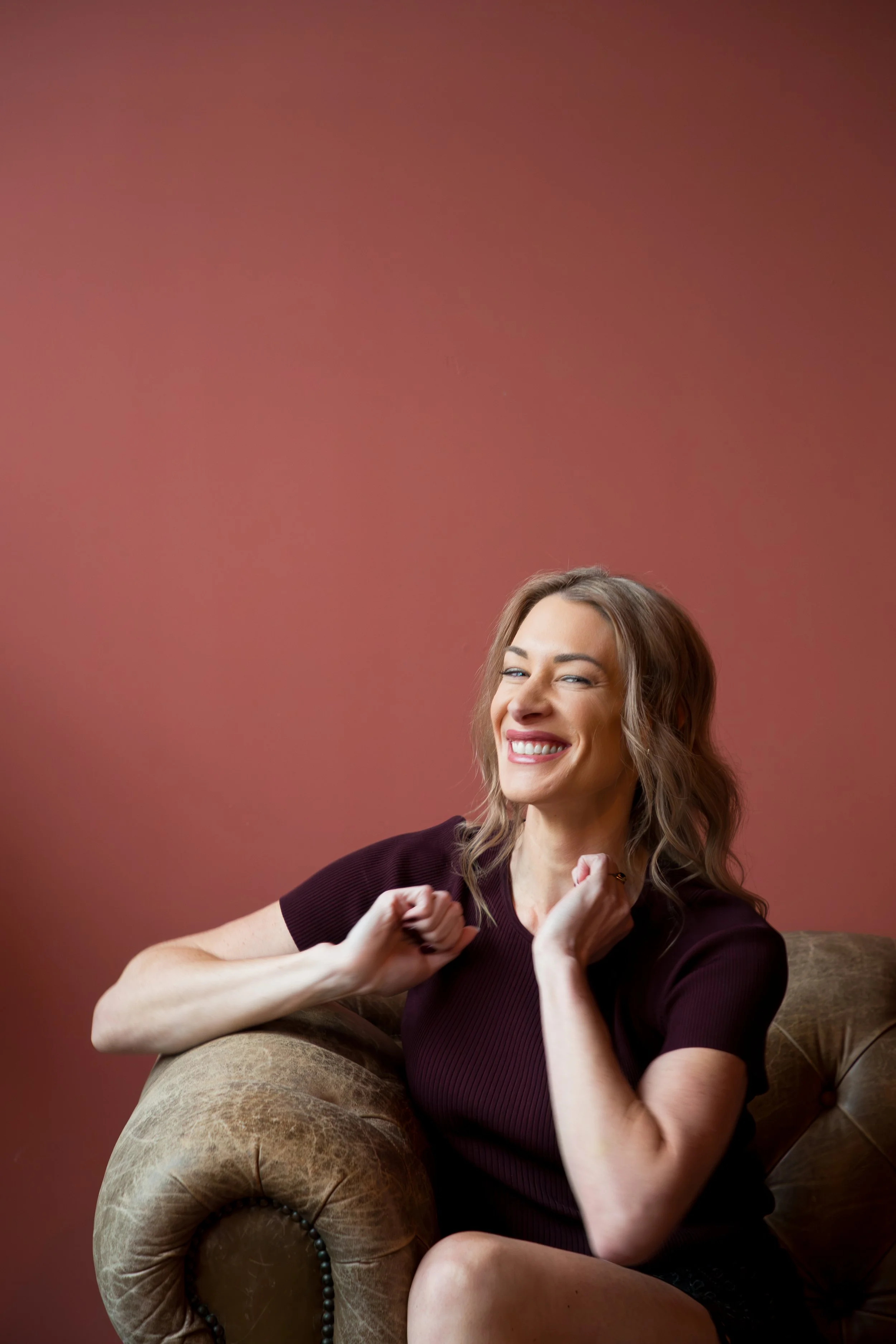 A woman with wavy blonde hair smiling and posing with her fists clenched in a playful fighting stance while sitting on a tan velvet armchair against a plain, reddish-brown background.
