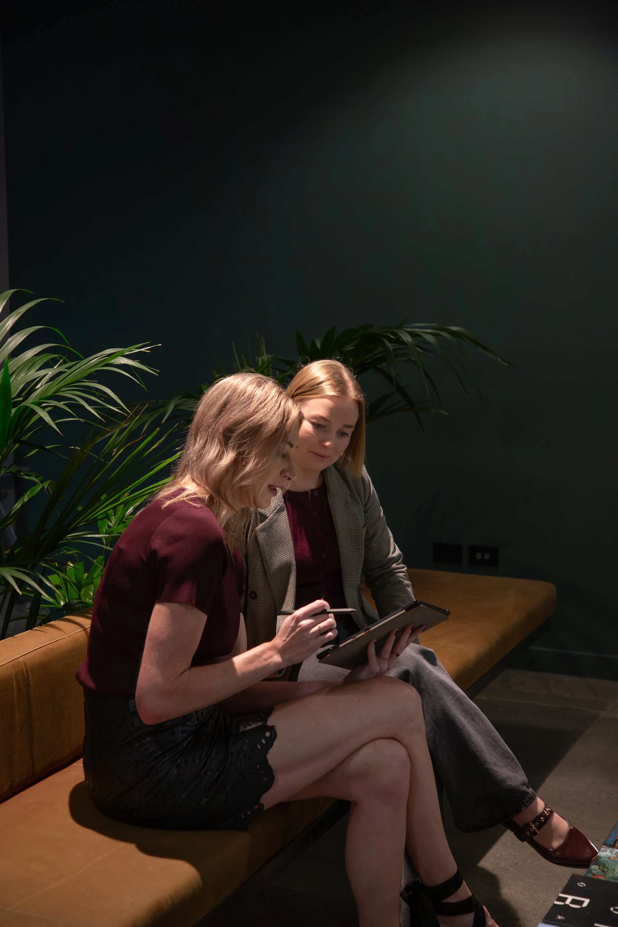Two women sitting on a brown velvet bench, looking at a tablet, with green plants behind them, in a dimly lit room.