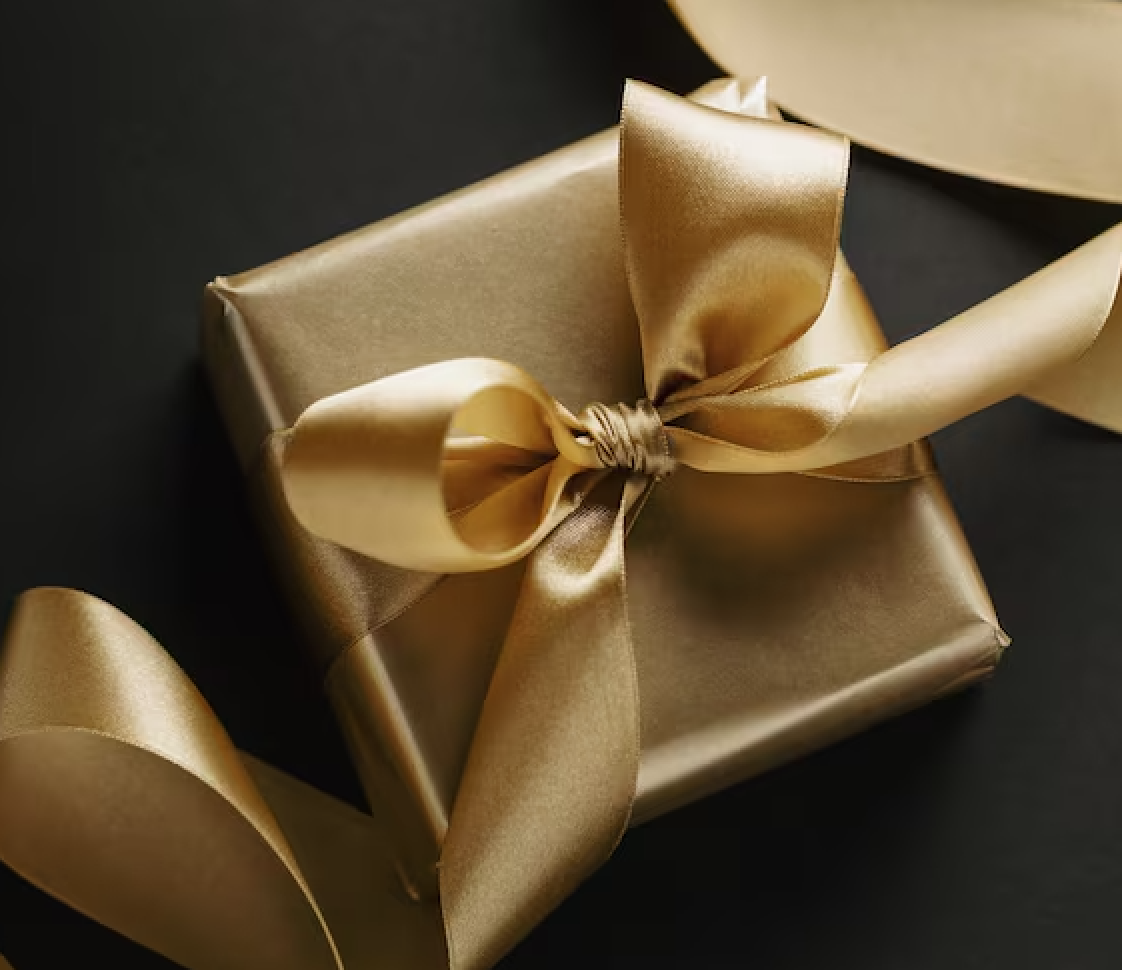 A gold wrapped gift box with a matching gold satin ribbon tied in a bow, resting on a black surface.