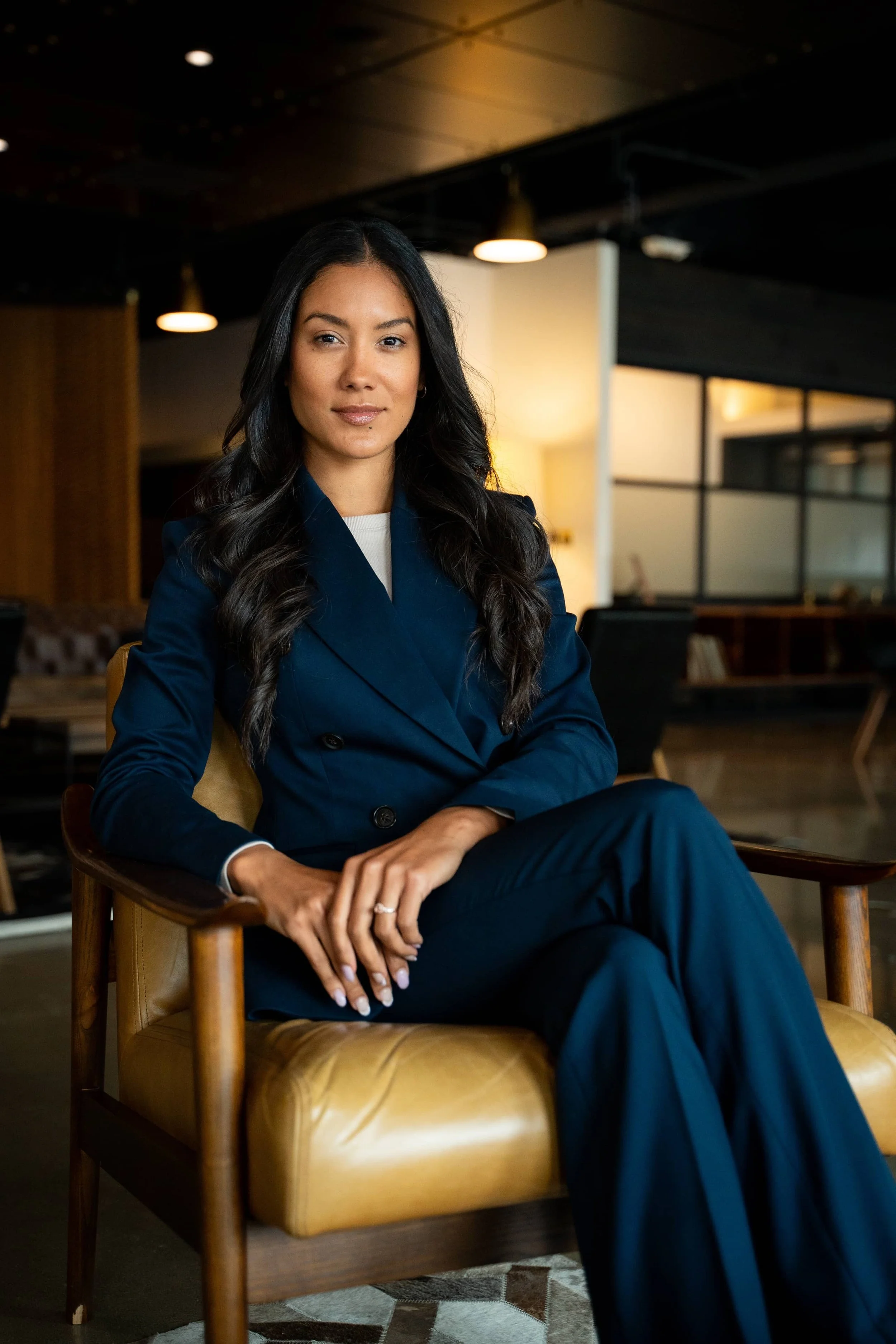 A woman with long black hair sitting in a wooden chair in a modern, dimly lit office space. She is wearing a navy blue blazer and looks confident.