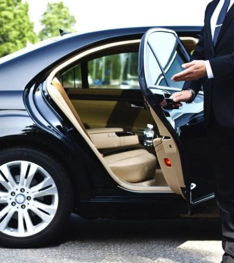 A person in a suit opening a rear door of a black luxury car, revealing a beige leather interior with a water bottle on the seat.