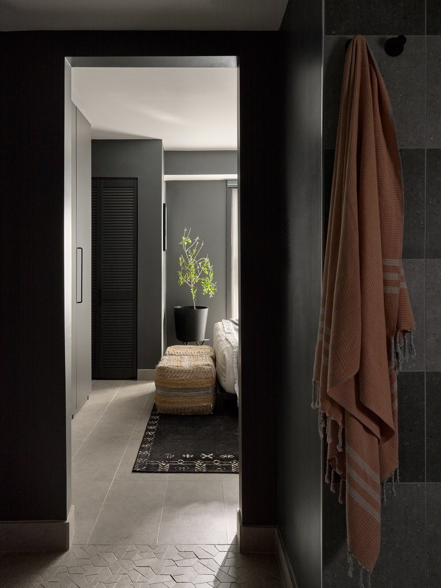 View of a modern, minimalistic hallway leading into a room with a potted plant, sofa, and a black cabinet. Clothes are hanging on a hook on the wall to the right, and a black patterned rug runs along the floor.