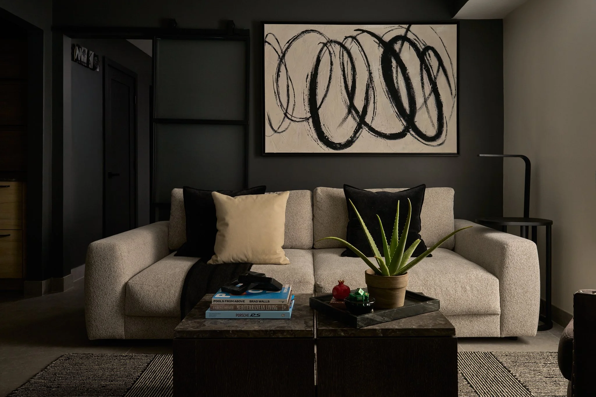 A living room with a beige sofa, black and beige pillows, a black marble coffee table, a green potted succulent, and a large abstract black and white painting on a dark gray wall.
