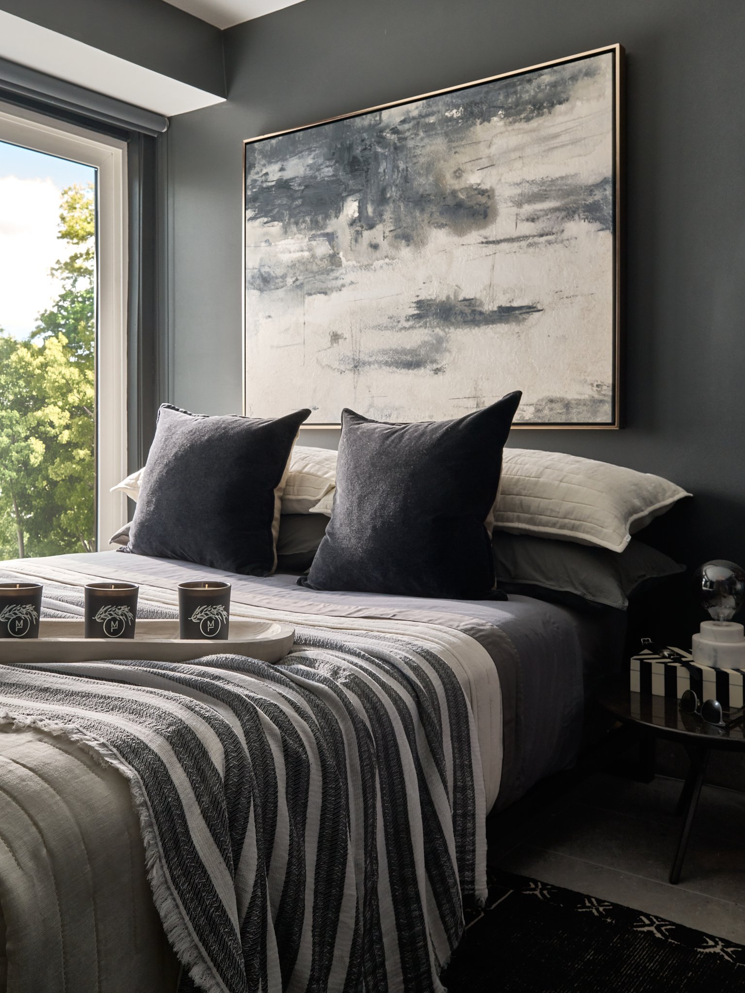 A cozy bedroom with grey and white striped bedding, black throw pillows, and a large abstract painting on a dark grey wall. There is a window showing trees outside, a tray with three candles on the bed, and a small black side table with white and black boxes and scented candles.