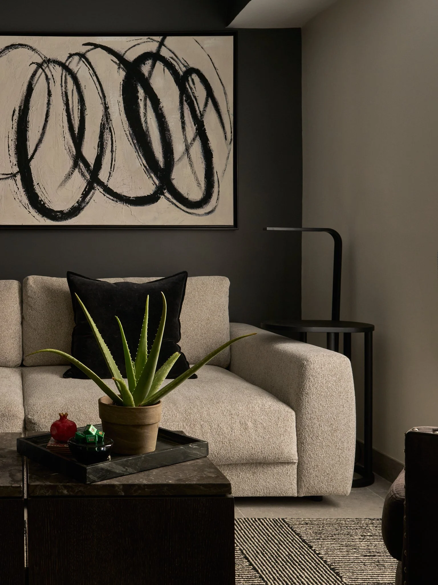 Living room with a beige sofa, black and beige pillows, a black marble coffee table with a potted aloe vera plant, a black tray with decorative items, a large abstract black and white painting on the wall, a black side table with a lamp, and a rug with black and white stripes.