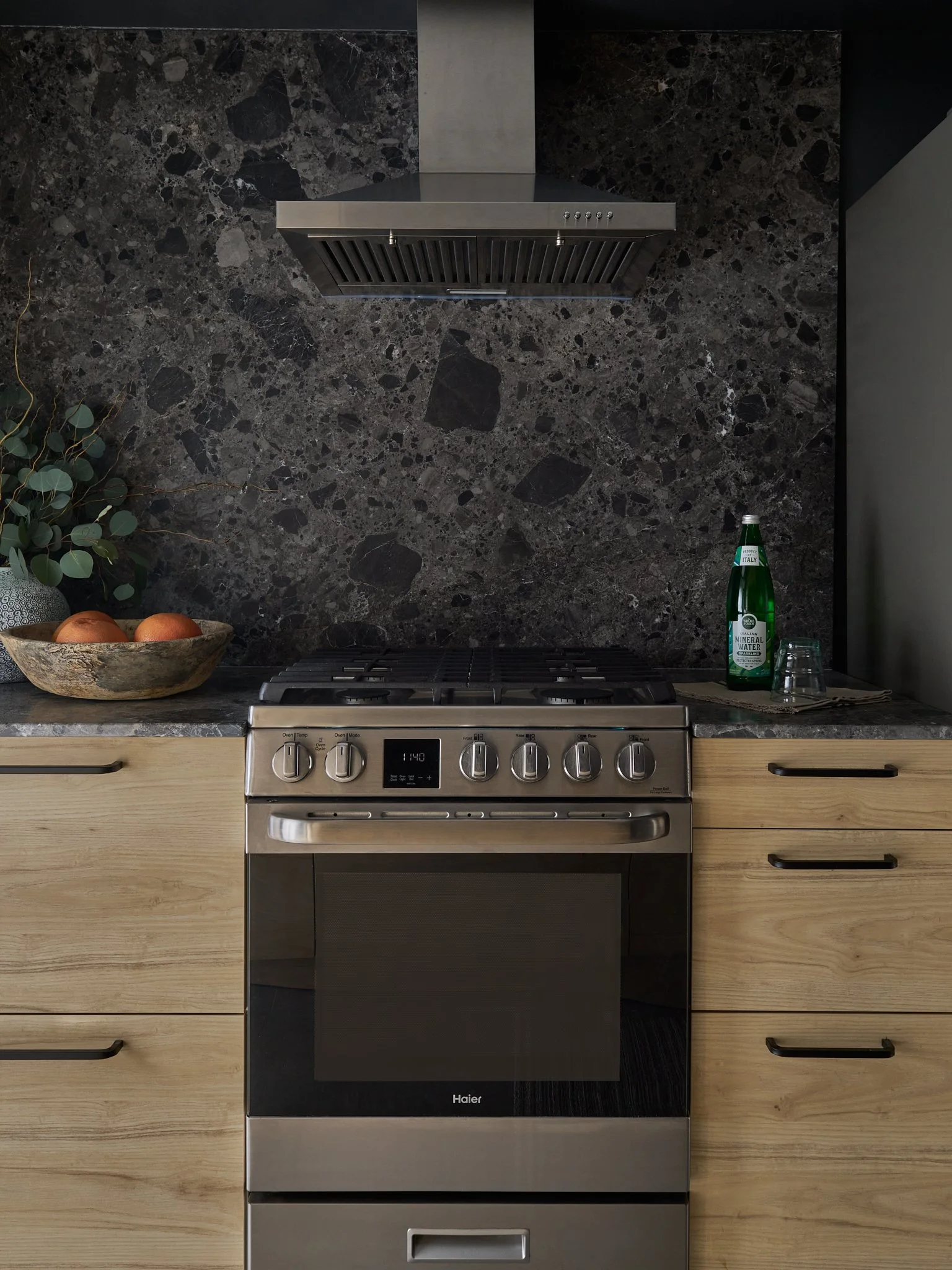 A modern kitchen with a black granite backsplash, a stainless steel range hood, and a stainless steel oven with a digital display. To the left, a bowl of eggs and a vase with greenery can be seen; on the right, there is a bottle of sparkling water and a glass on a napkin.